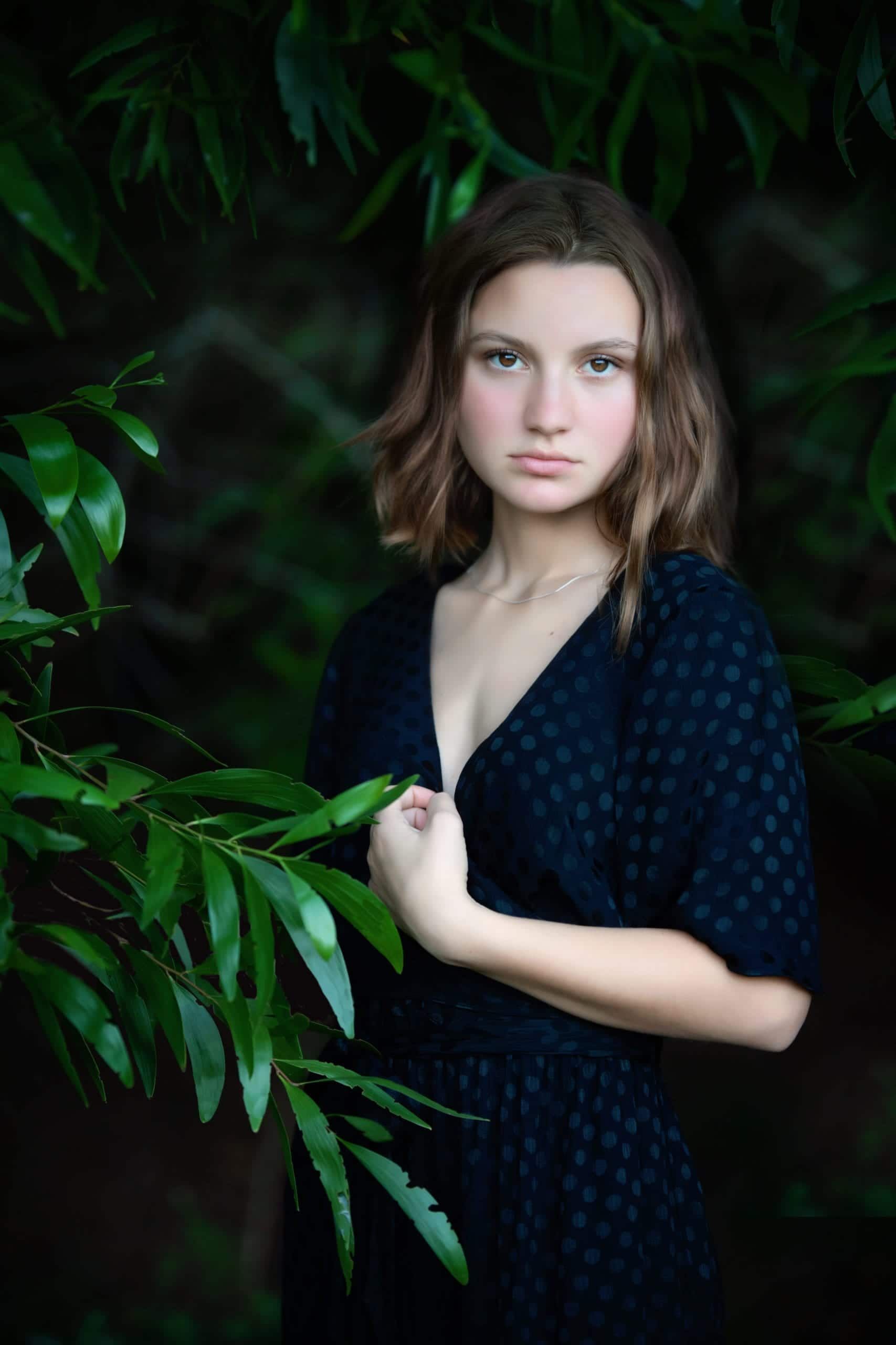 Young woman in a black dress poses in front of foliage, looking directly at the camera.
