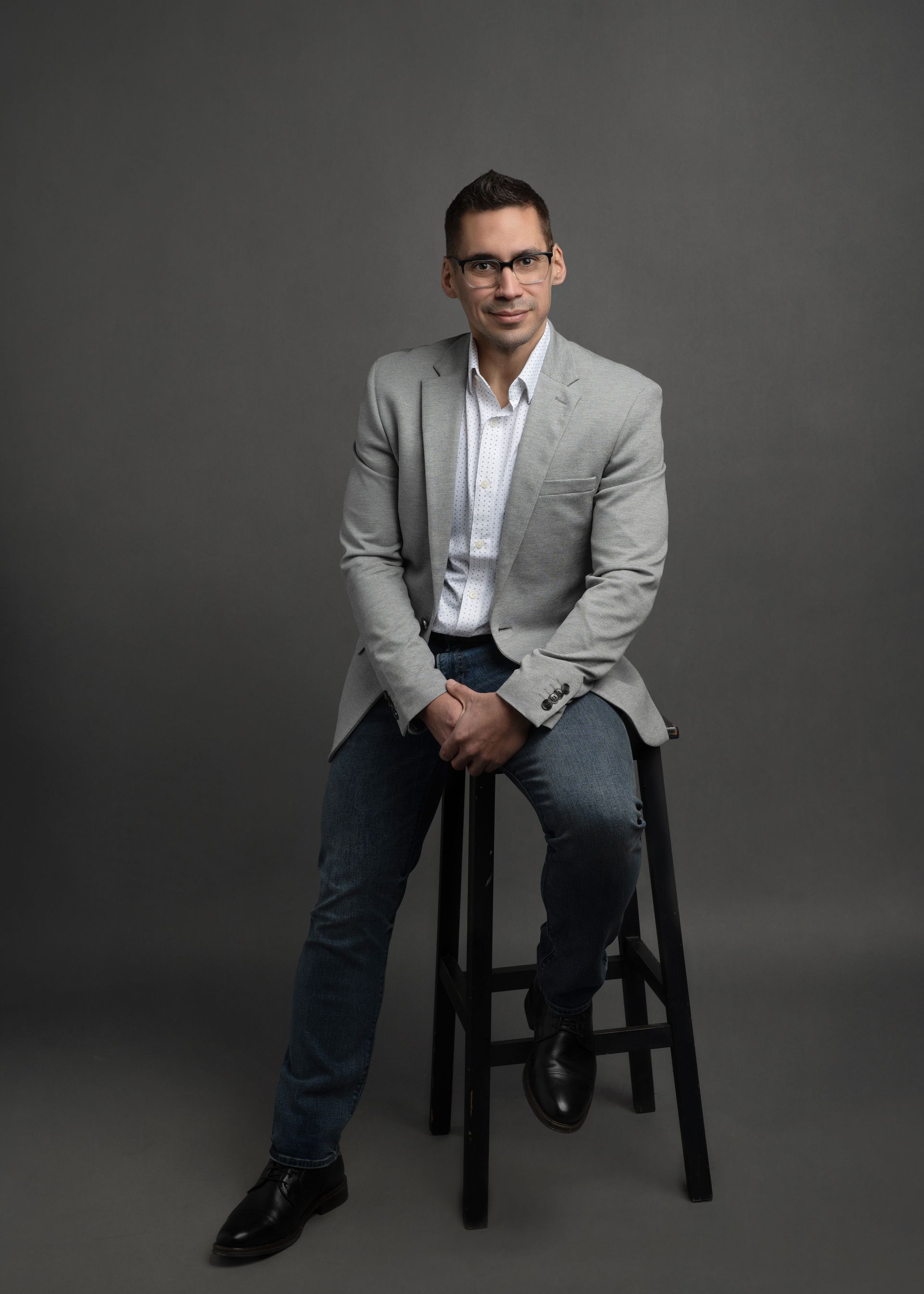 Man in blazer, jeans, and glasses sits on a stool against a gray backdrop, looking at the viewer.