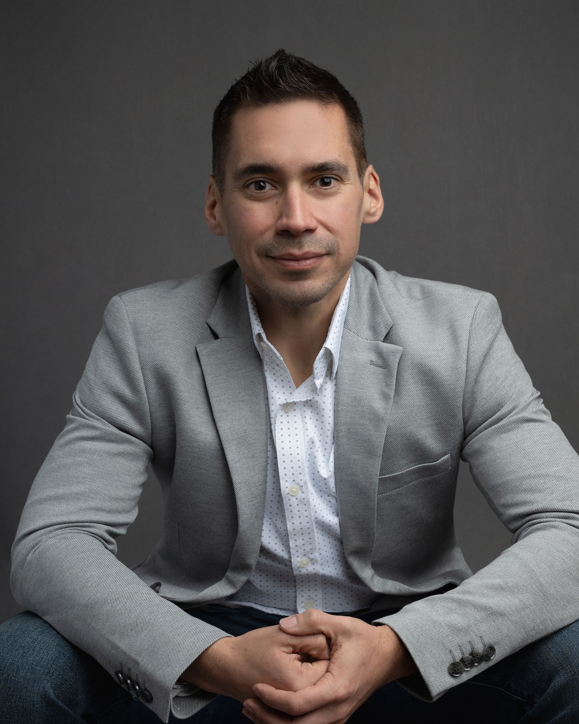 Man in light gray blazer, white shirt, and jeans sits looking forward, hands clasped; gray background.