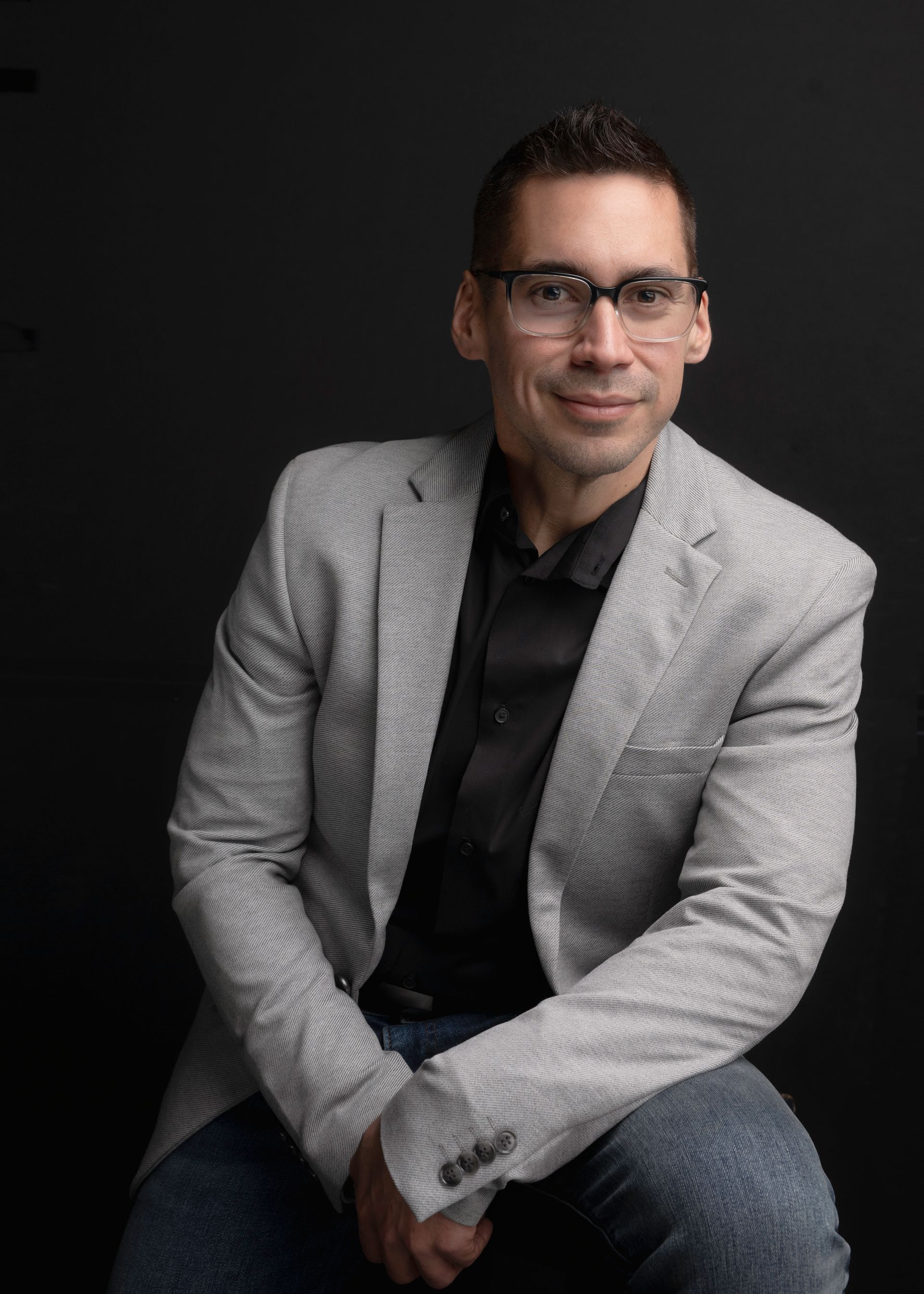 Man in glasses, light gray blazer, black shirt, jeans, smiles, dark background.