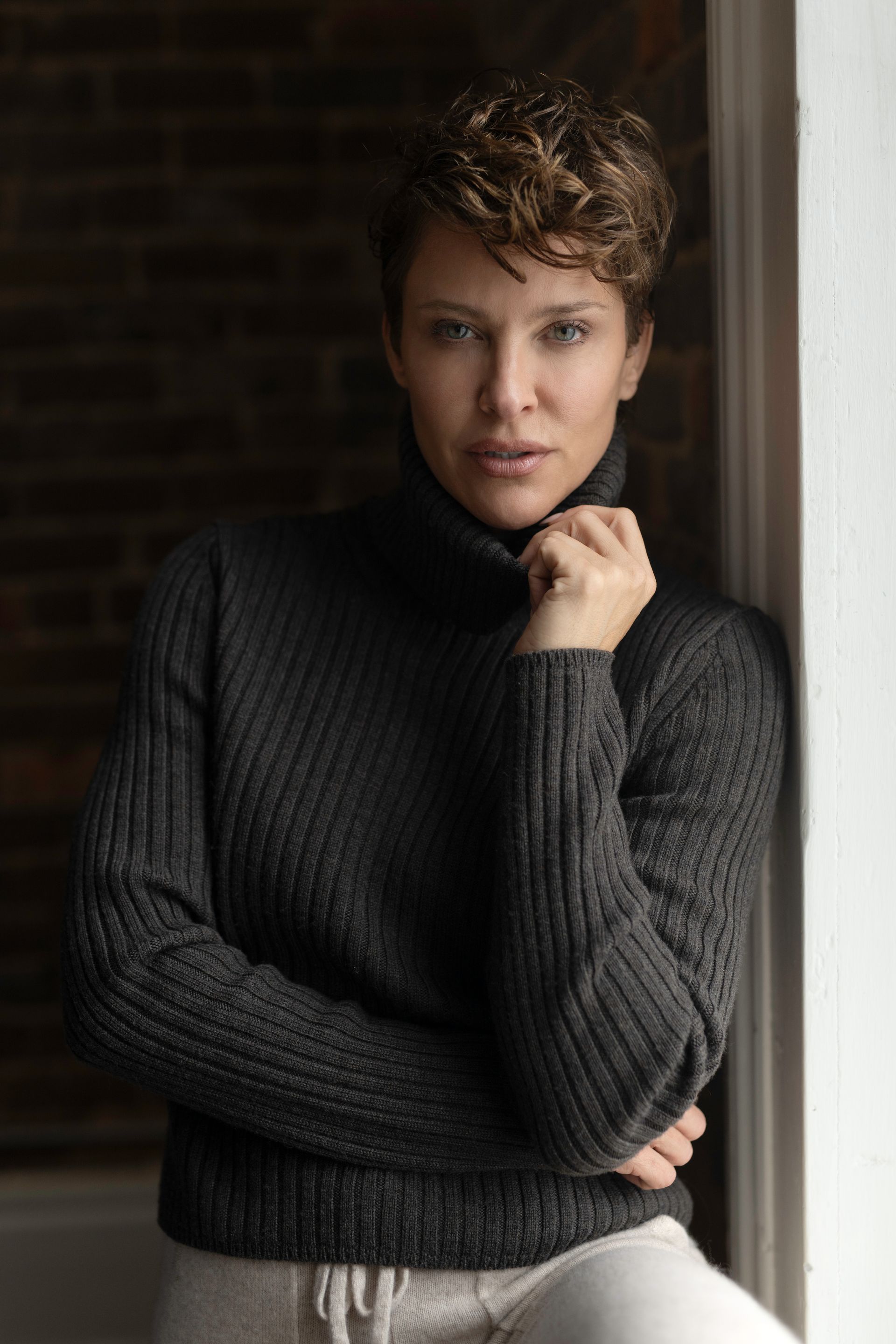 Woman with short, curly brown hair in a charcoal turtleneck, leans against a wall, looking at the camera.