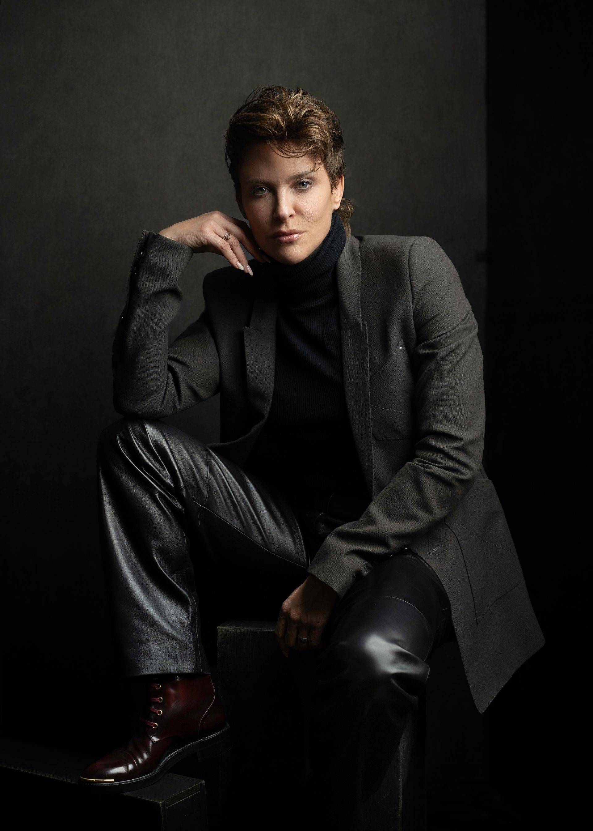 Woman in leather pants, blazer, and turtleneck, sitting with a hand on her face. Dark background.