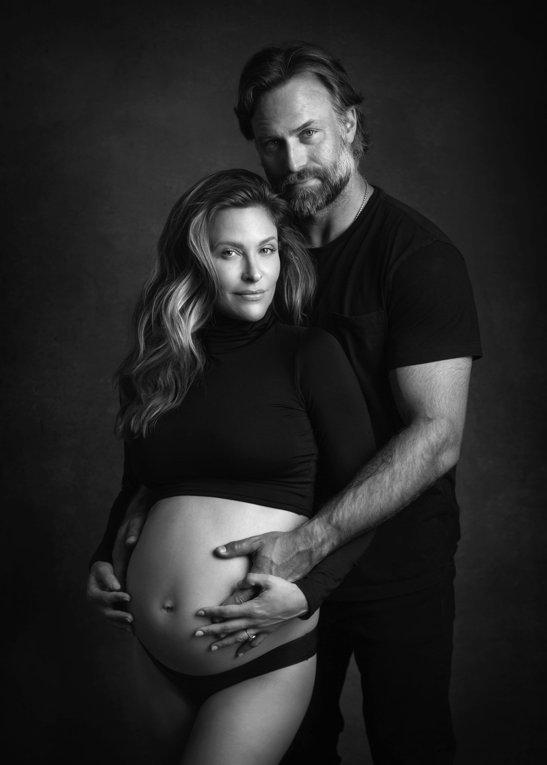 Pregnant woman and man in black attire, posing together in a studio.