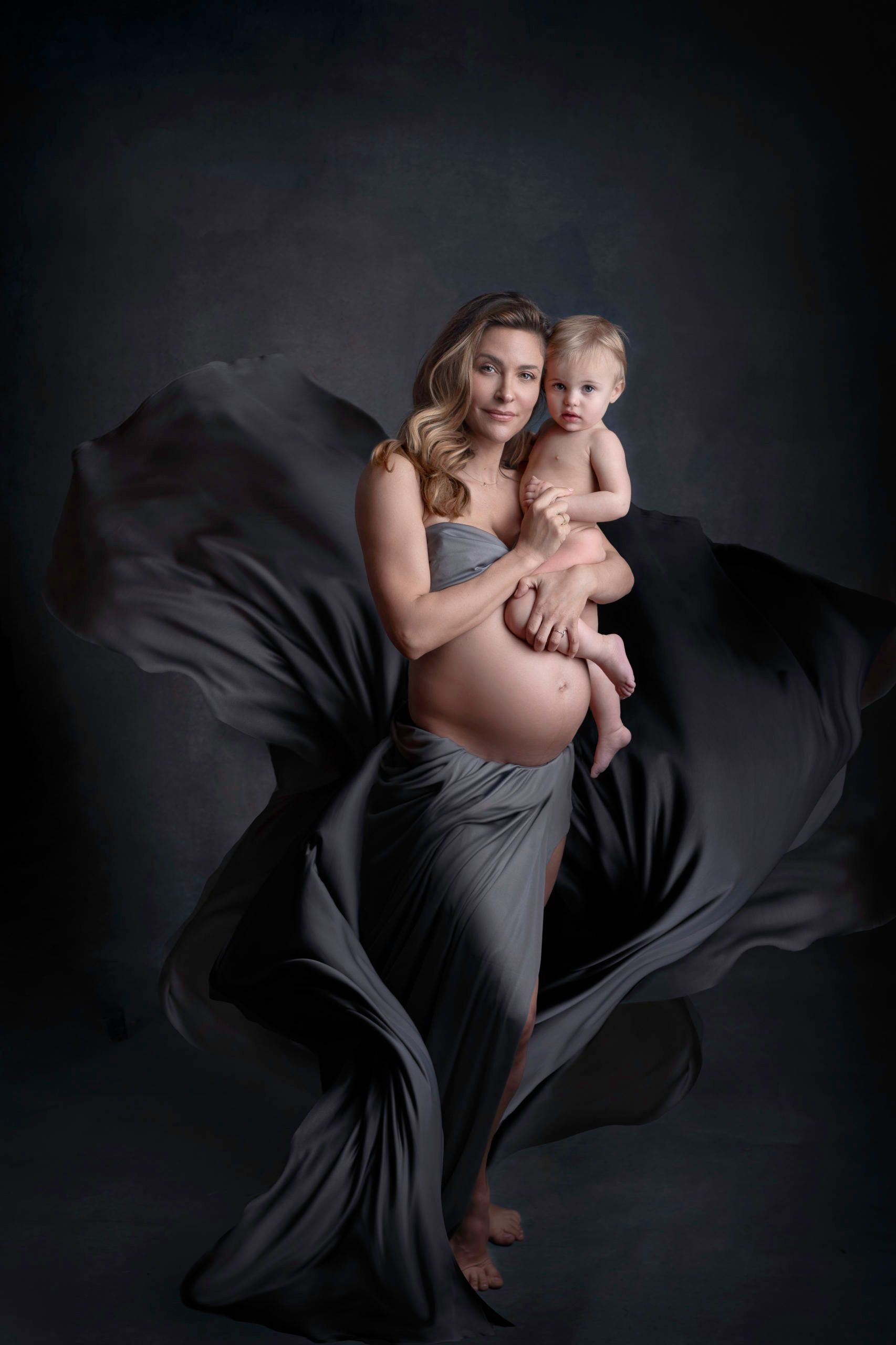 Pregnant woman holding baby; draped in gray fabric, dark background.