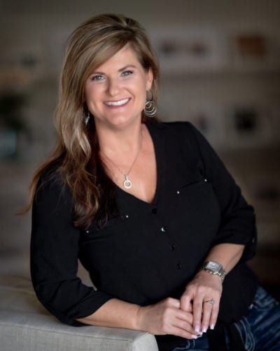 Woman with blonde hair smiles, wearing black top, leaning on couch.