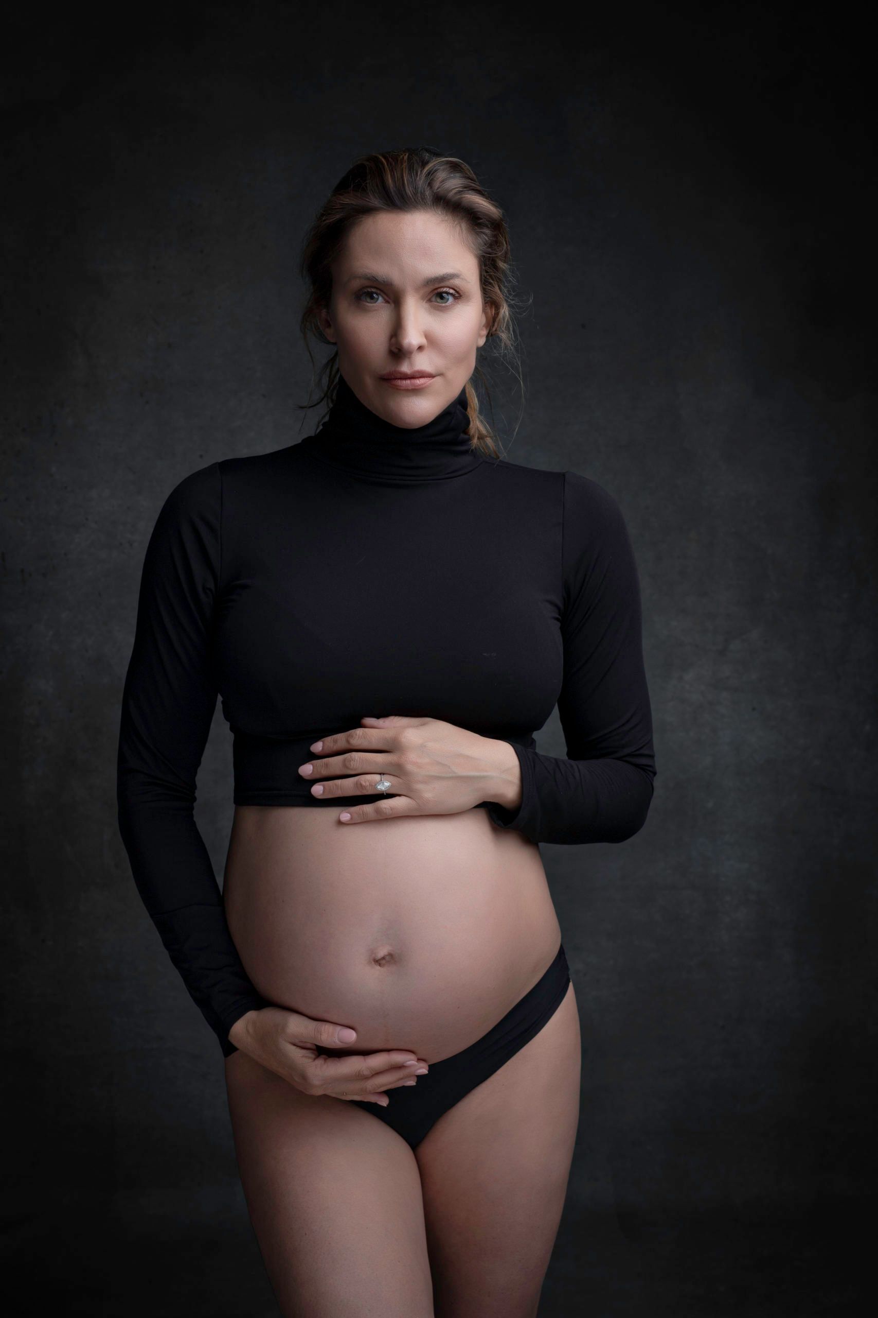 Pregnant woman in black turtleneck and underwear, hands on belly, against dark background.