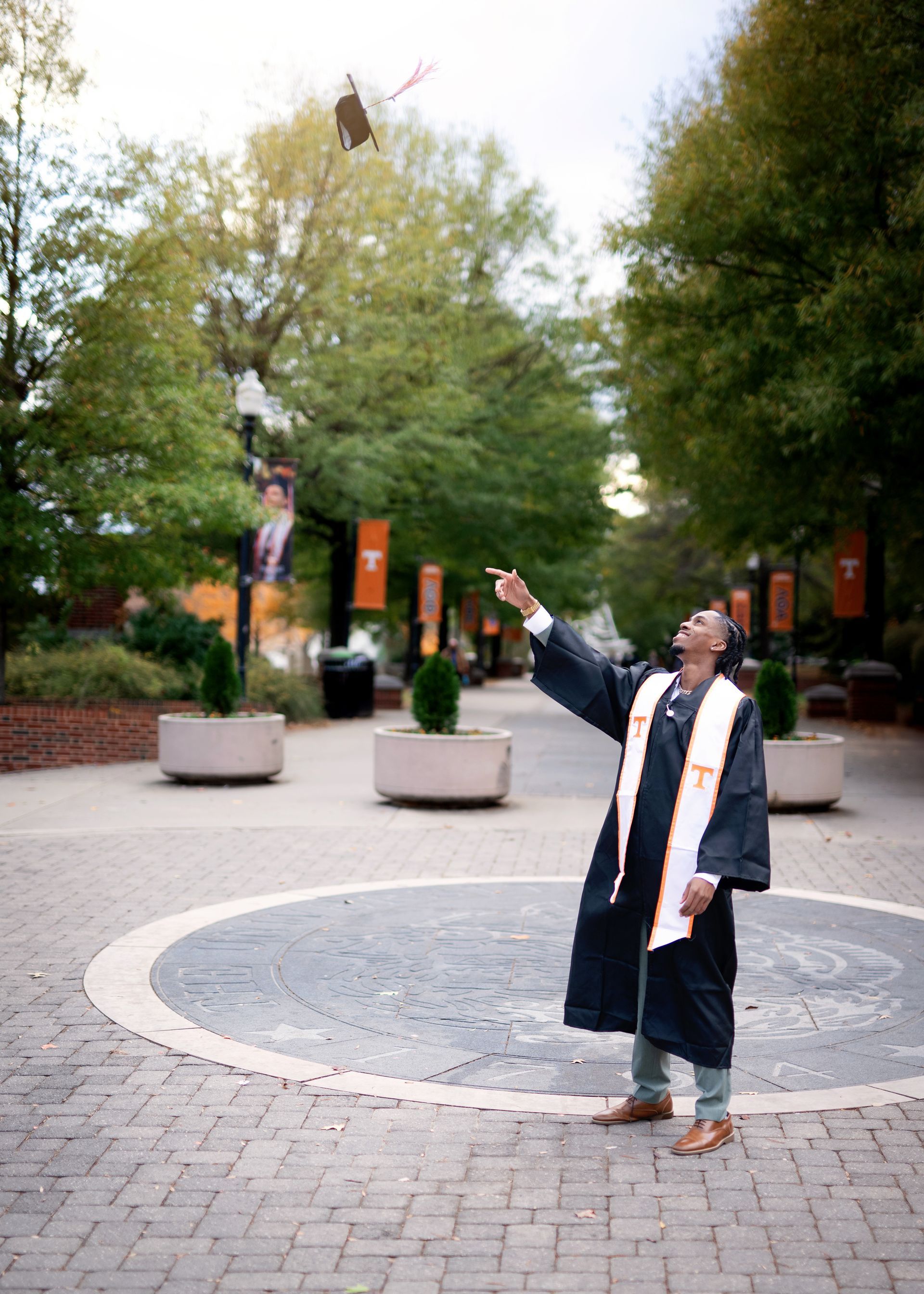 A graduate in a gown throws their cap in the air on a brick pathway lined with trees.