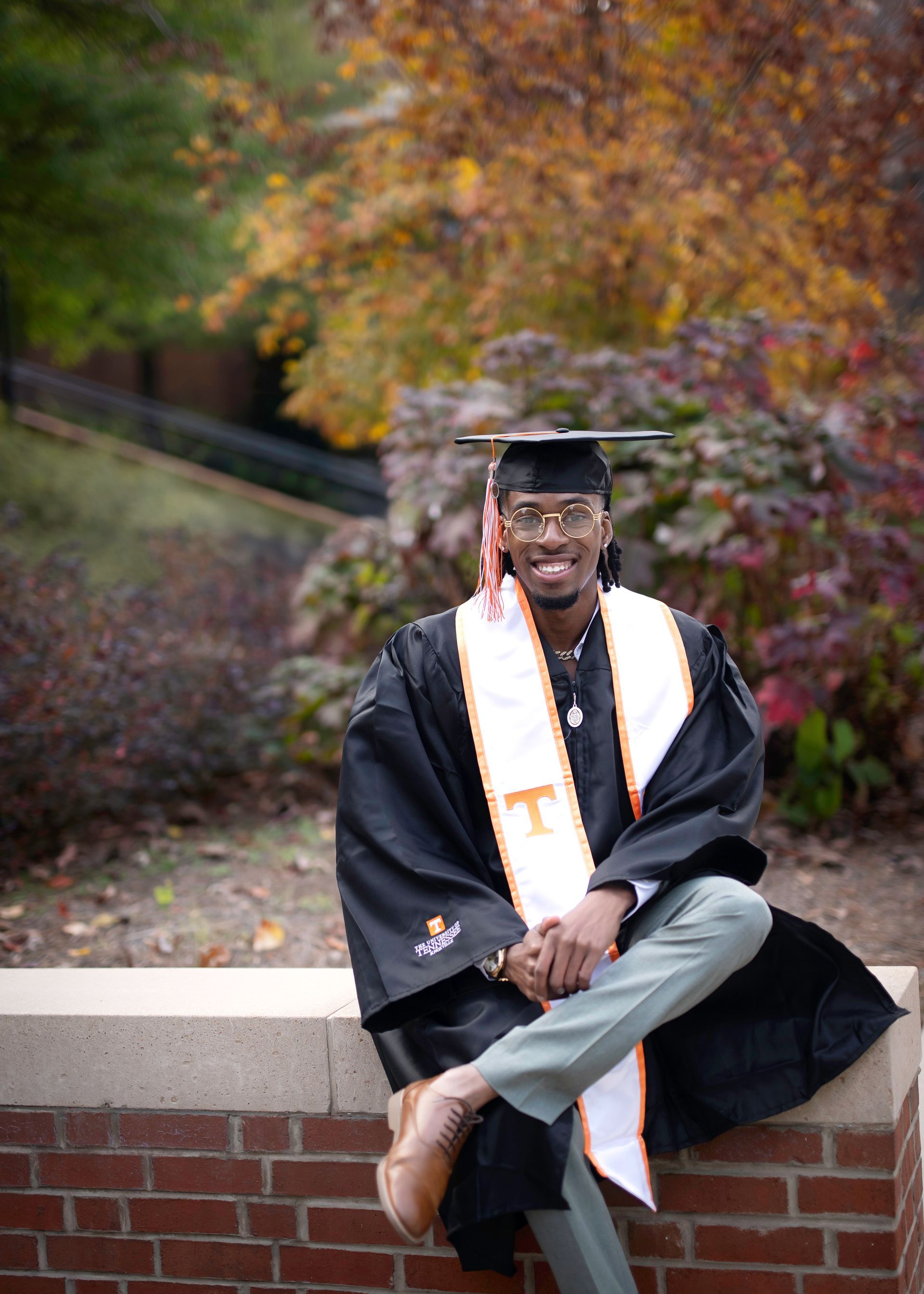 A smiling graduate in cap and gown, seated outdoors on a brick wall, fall foliage in background.
