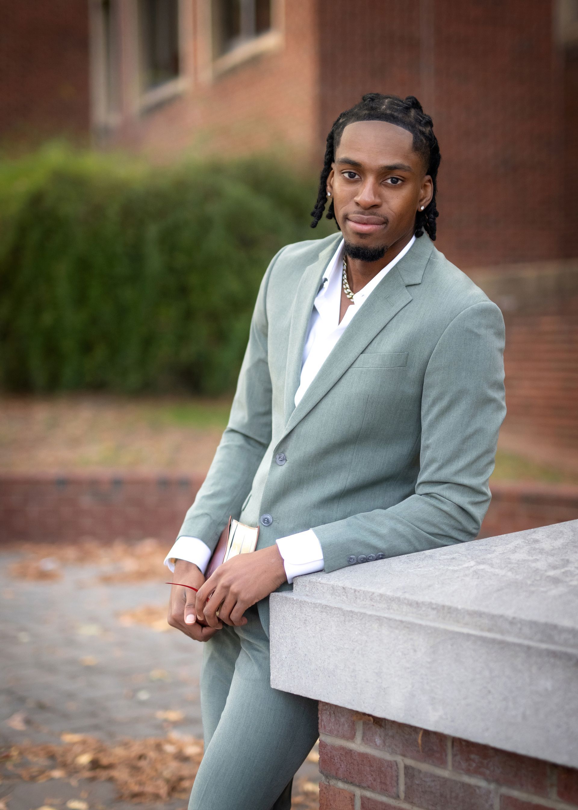 Man in green suit leaning on brick wall, holding a book, looking at the camera.