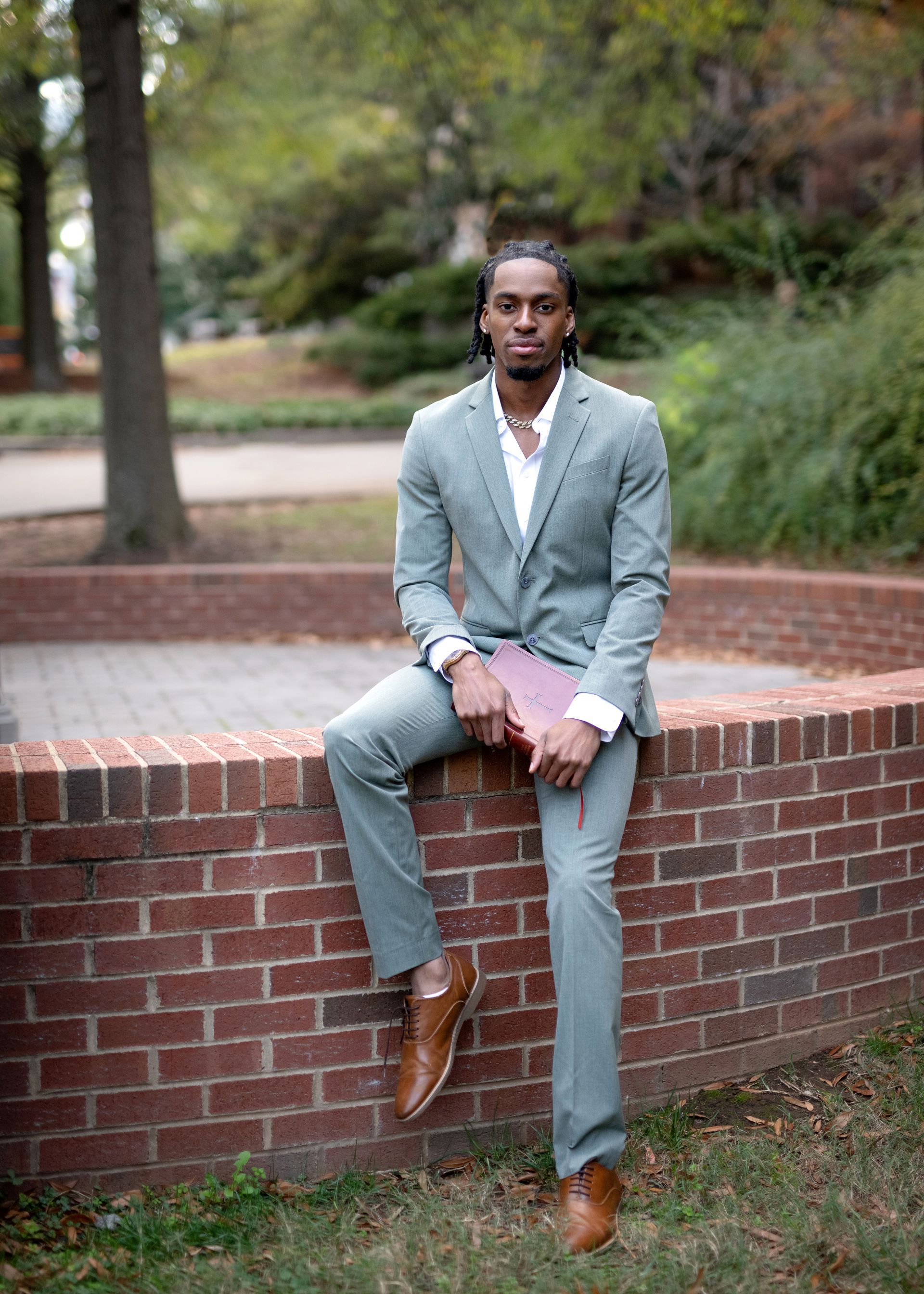 Man in green suit sitting on brick wall, holding book, outside.