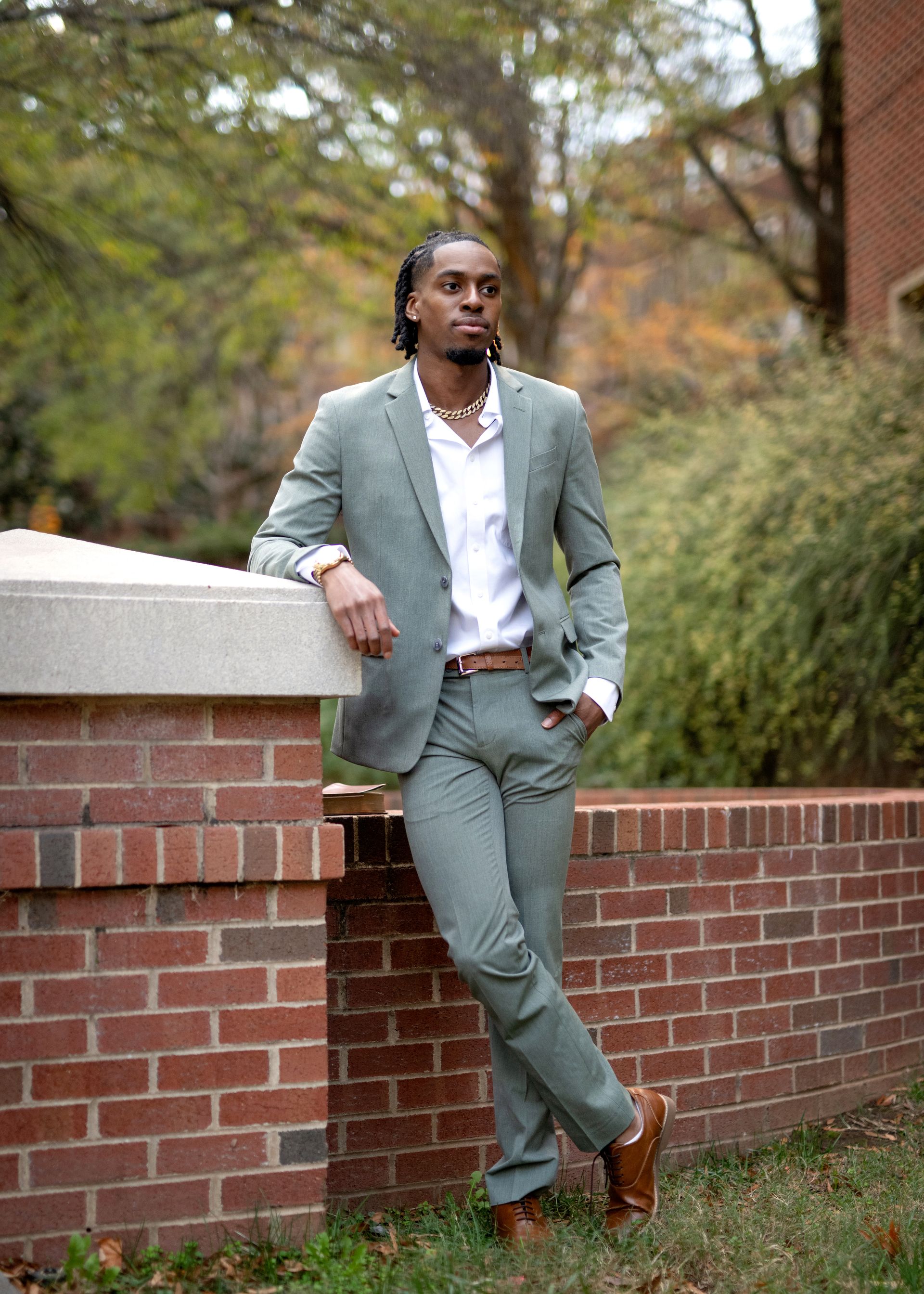 Man in green suit leans against a brick wall, smiling. Outdoors, trees in background.