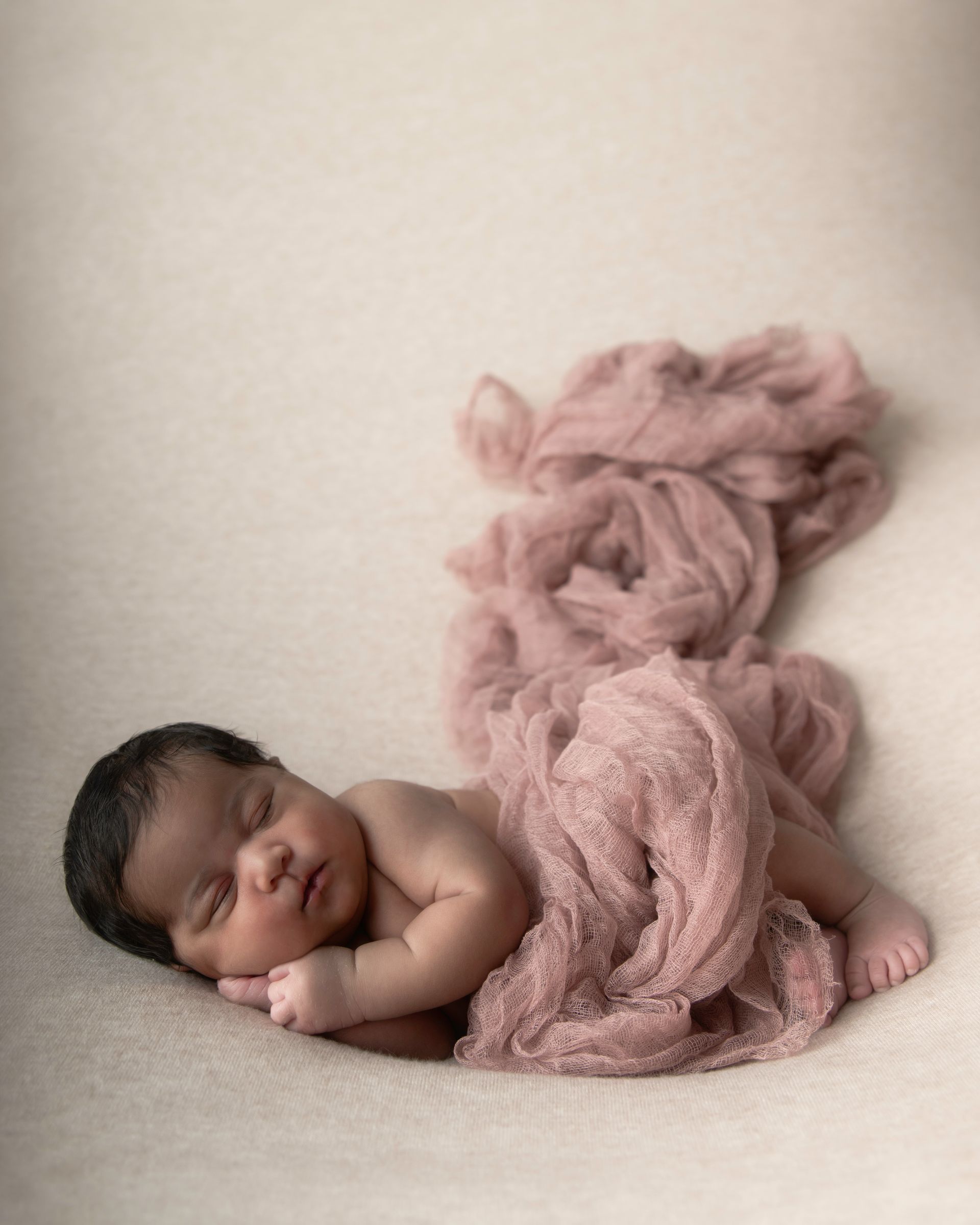 Sleeping newborn, dark hair, wrapped in pink fabric, on a neutral backdrop.