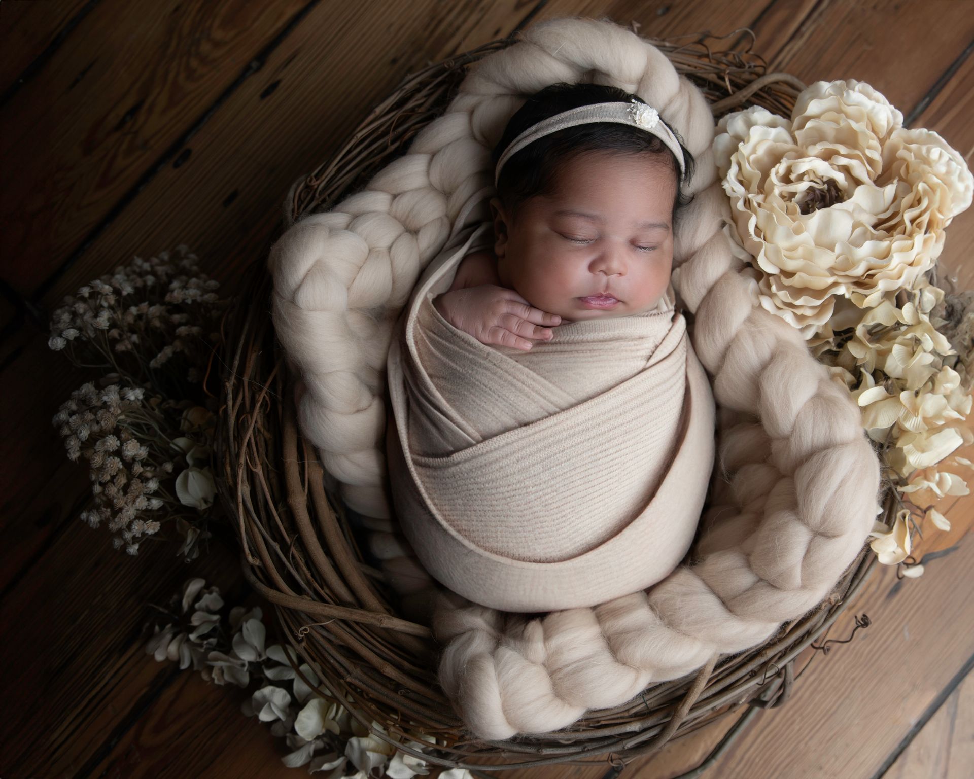 Newborn baby wrapped in cream-colored fabric, nestled in a woven nest, with floral accents.