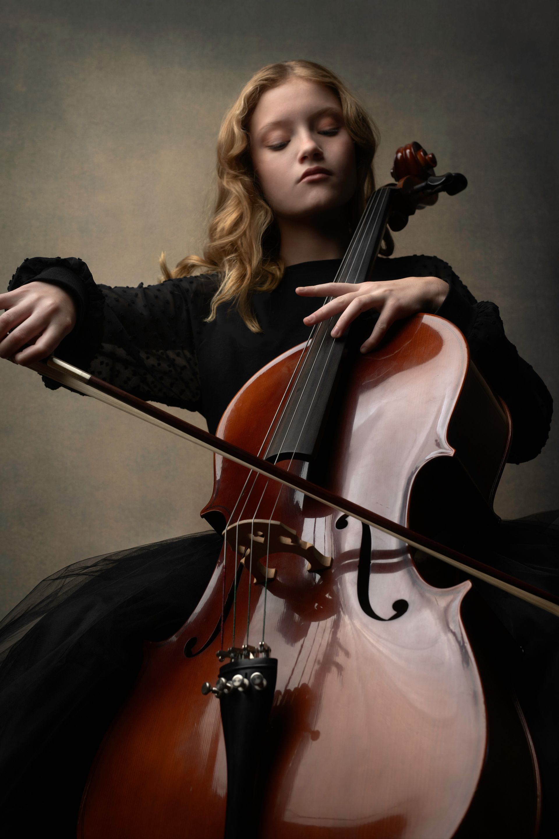 Young woman playing a cello, eyes closed, wearing black dress, against a muted background.