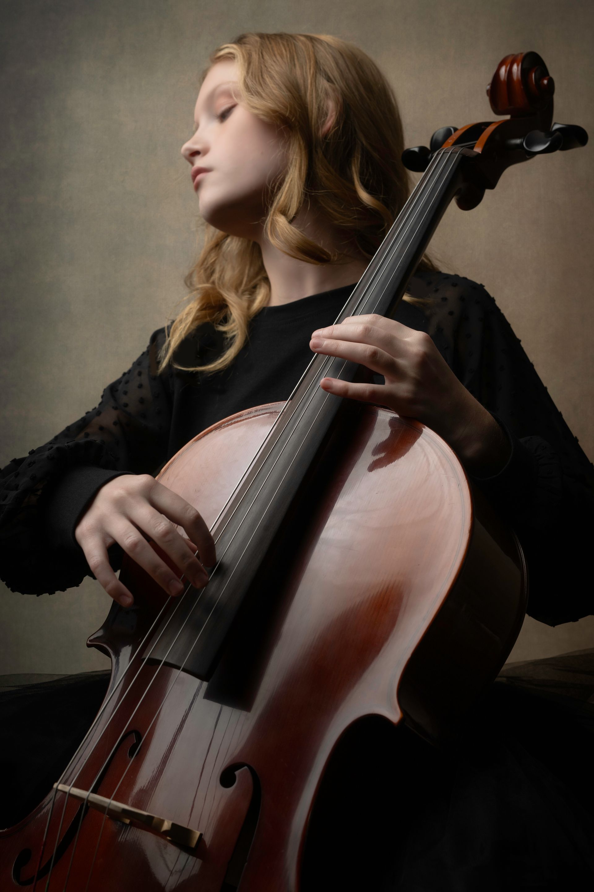 Young girl with long blonde hair plays the cello, eyes closed, wearing a black top.