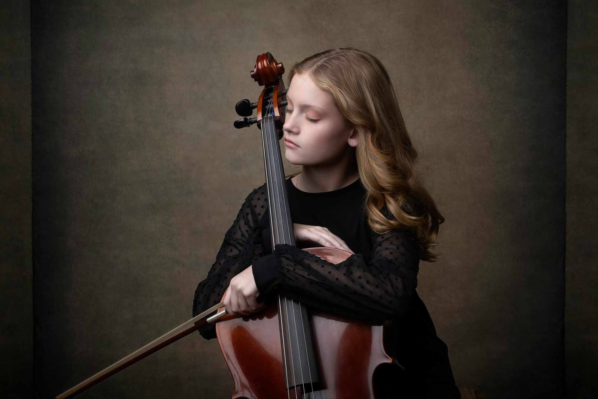 Young girl with cello, eyes closed, leaning on instrument, brown background.