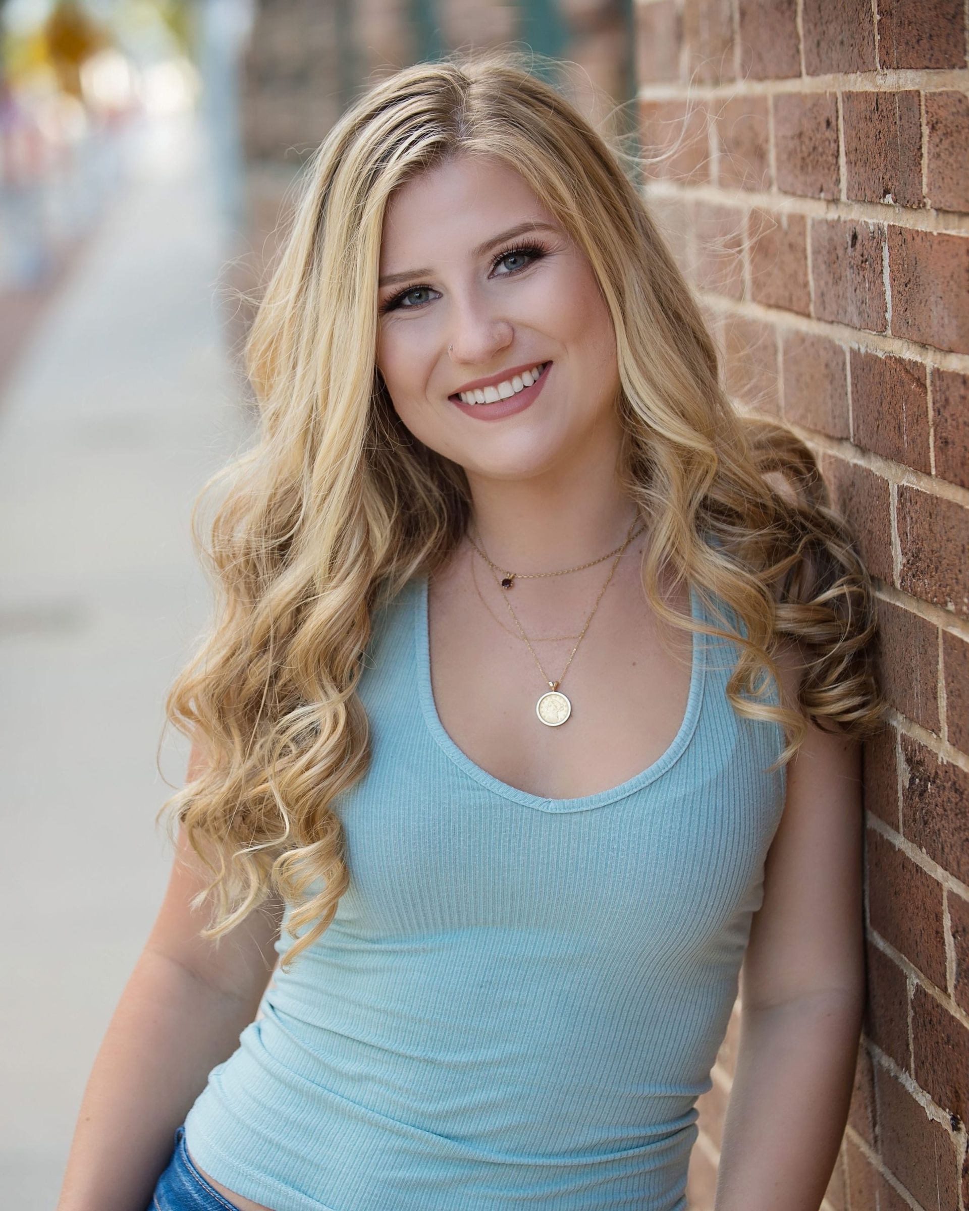 Blonde woman in a blue top leans against a brick wall, smiling.