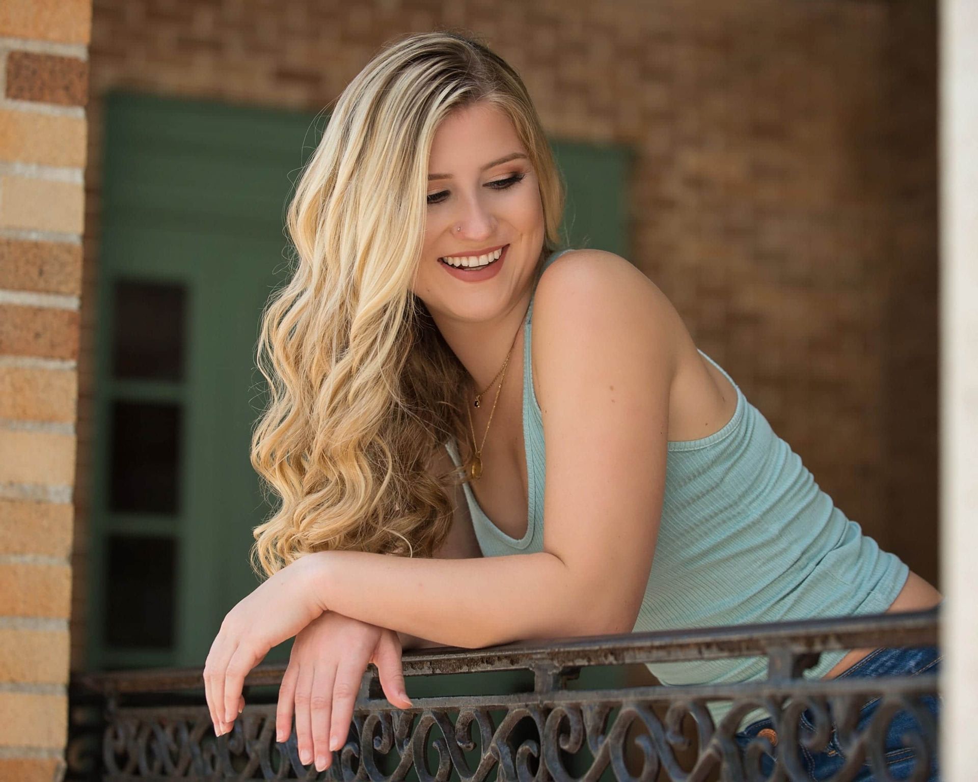 Blonde woman in a turquoise tank top smiles, leaning on a wrought iron balcony. Brick building background.