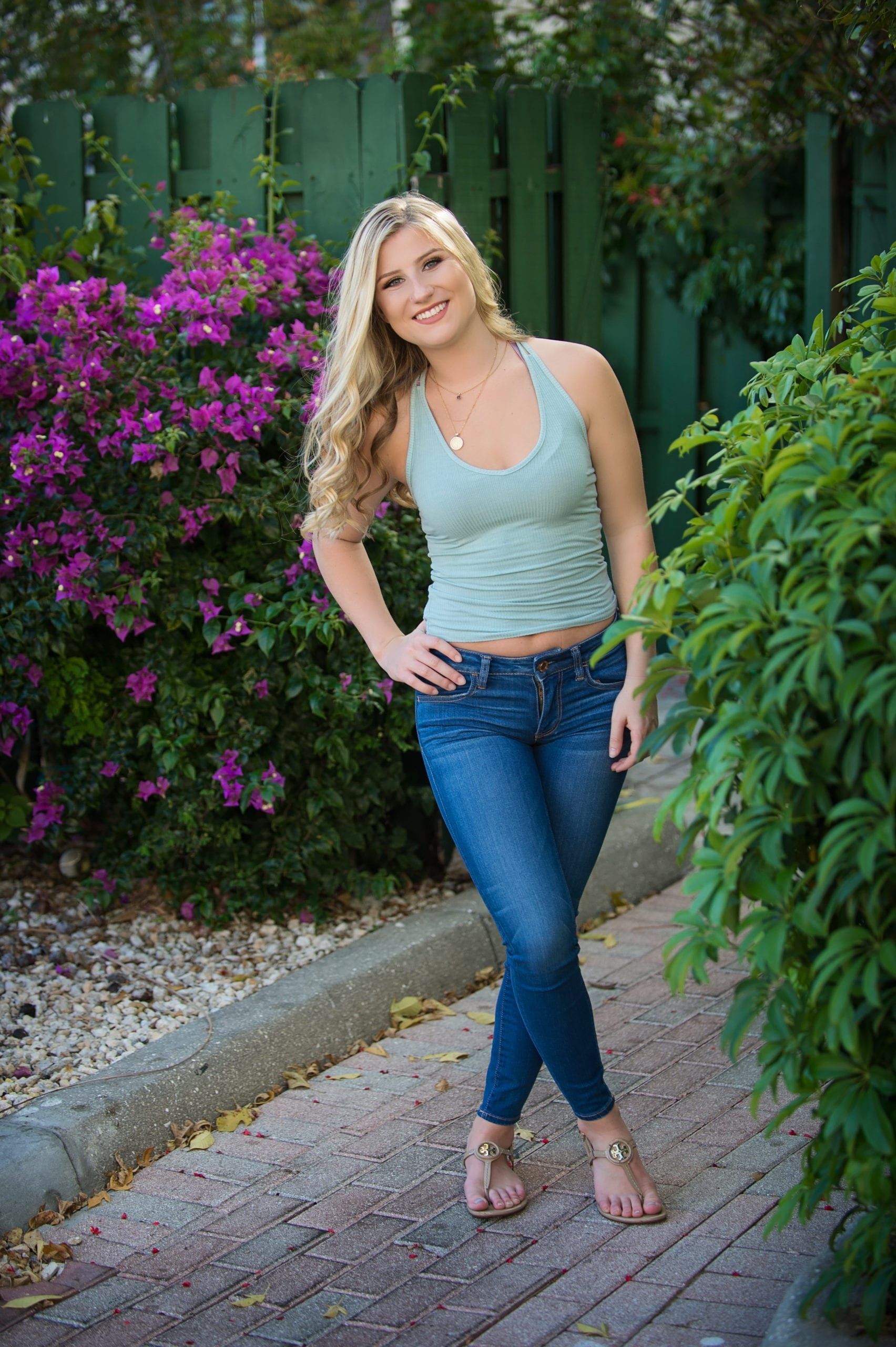 Blonde woman in jeans and green top posing outside near purple flowers and green foliage.