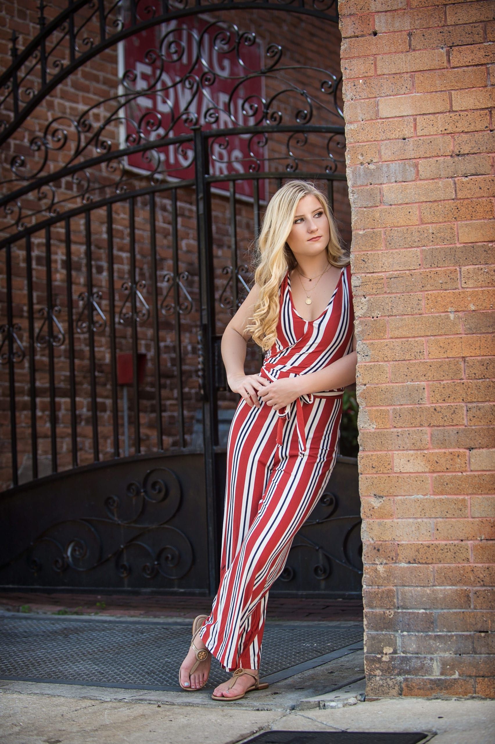 Blonde woman in red and white jumpsuit leans against a brick wall.  Ornate iron gate in background.