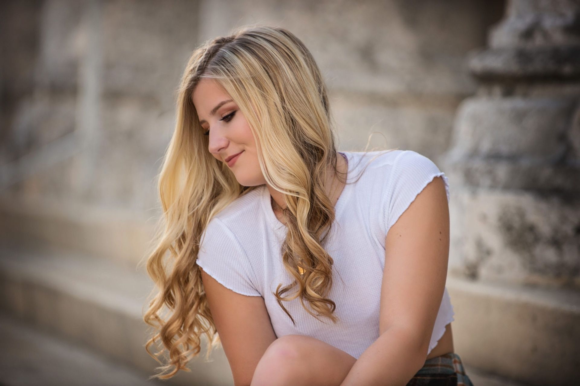 Blonde woman in white top smiles and looks down, posing on stone steps outside.