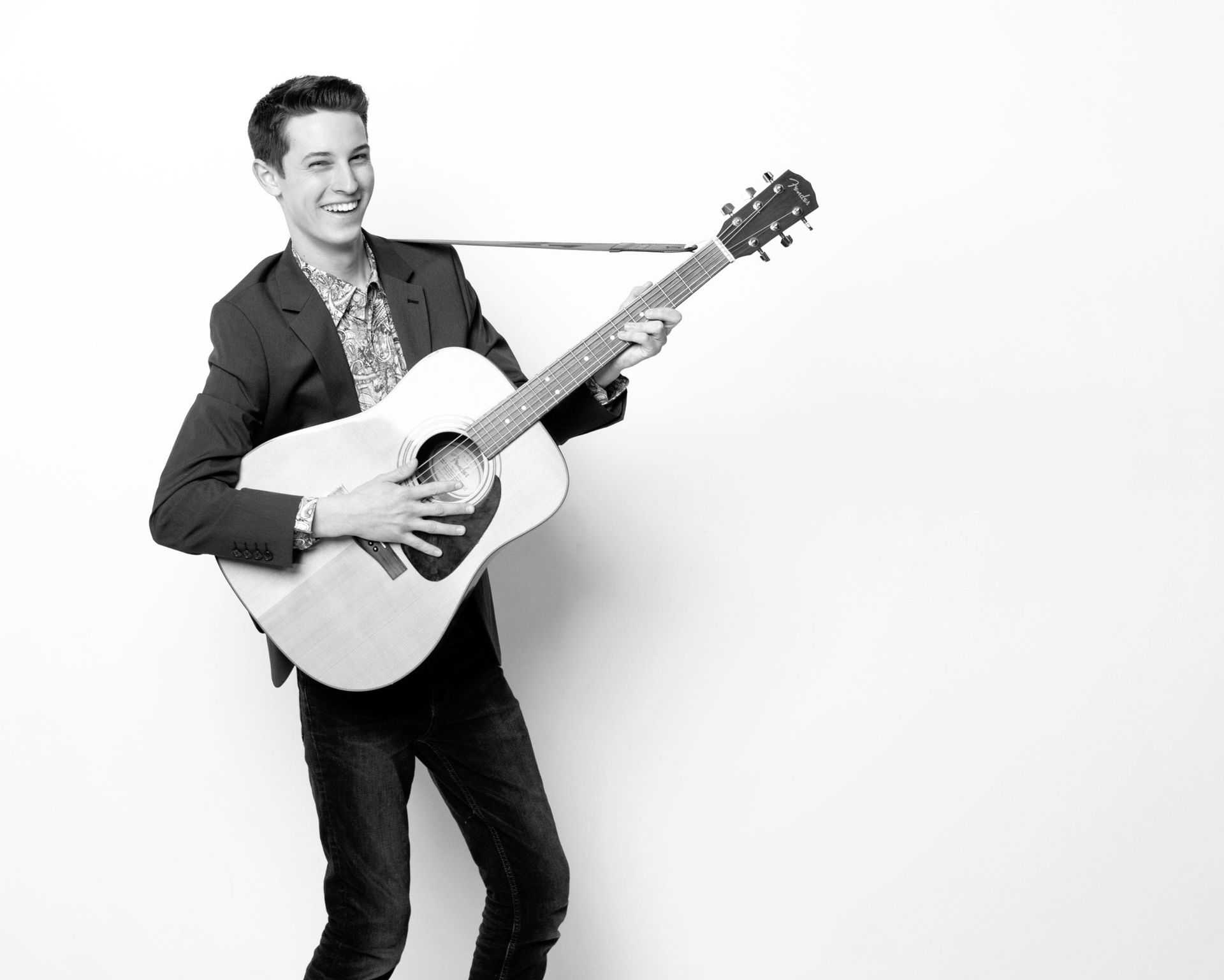 Man playing acoustic guitar, smiling, wearing suit jacket and patterned shirt, against white background.