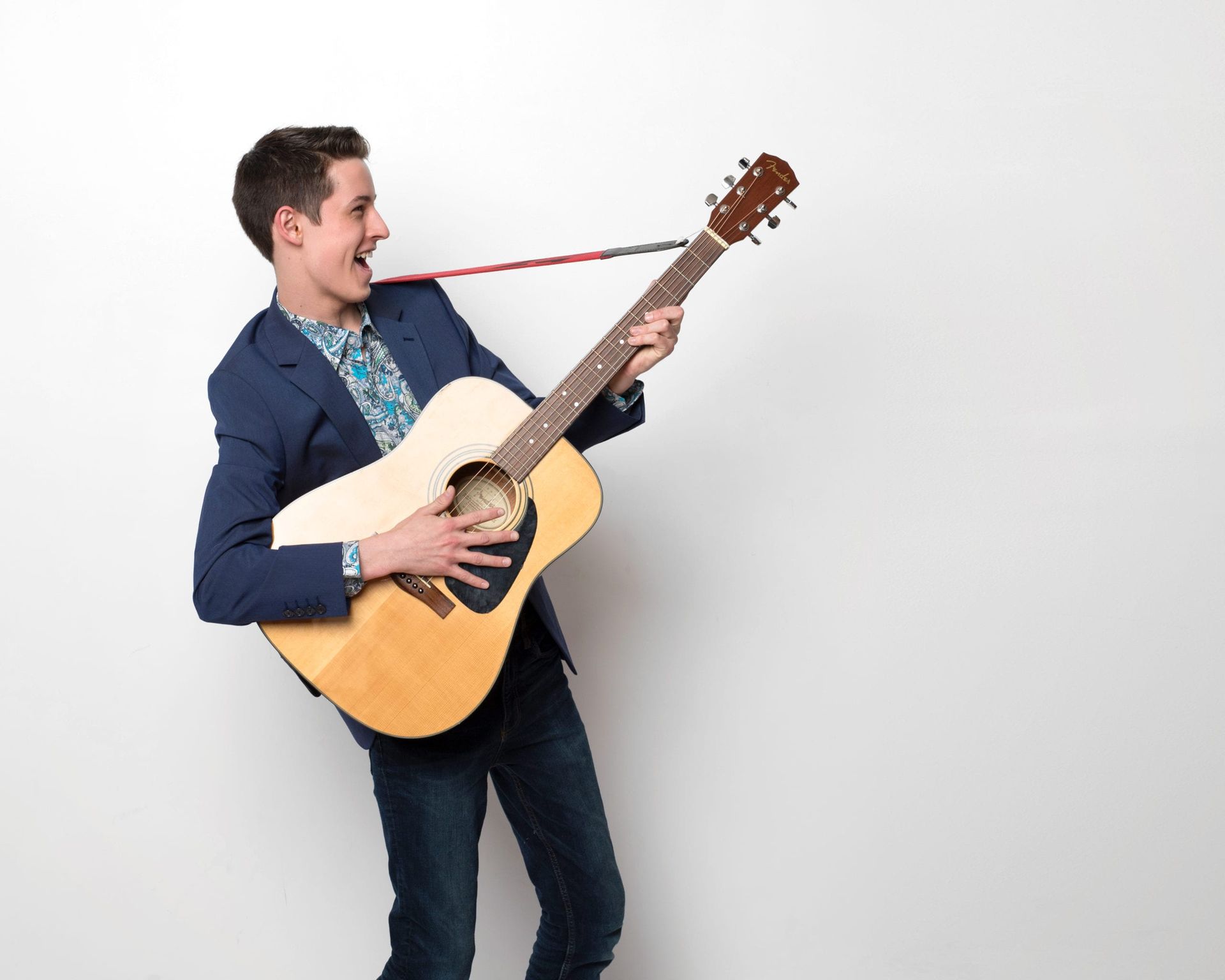 Young man in blue blazer playing acoustic guitar with tongue out.