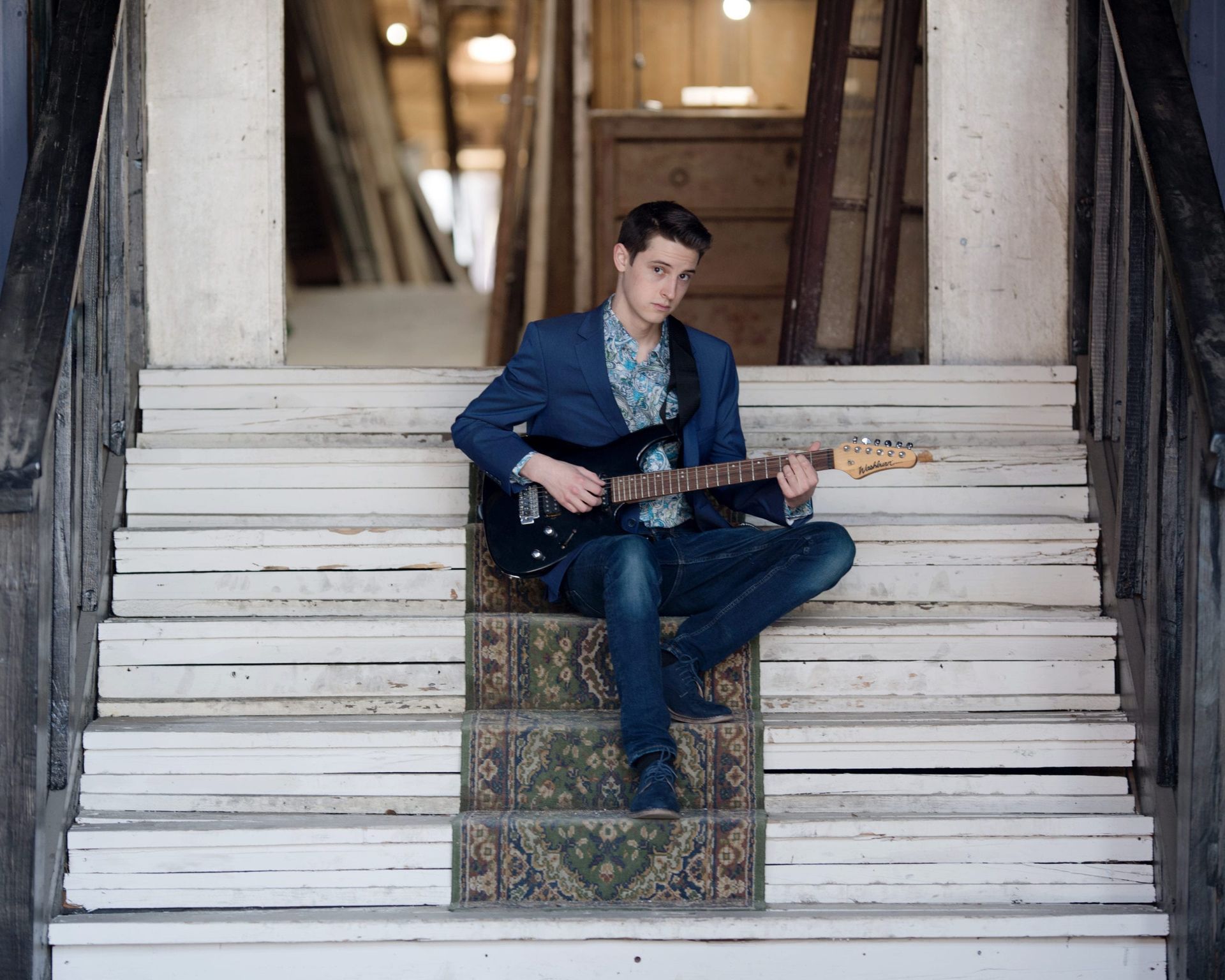 Young man in blue blazer playing guitar on old, weathered stairs.
