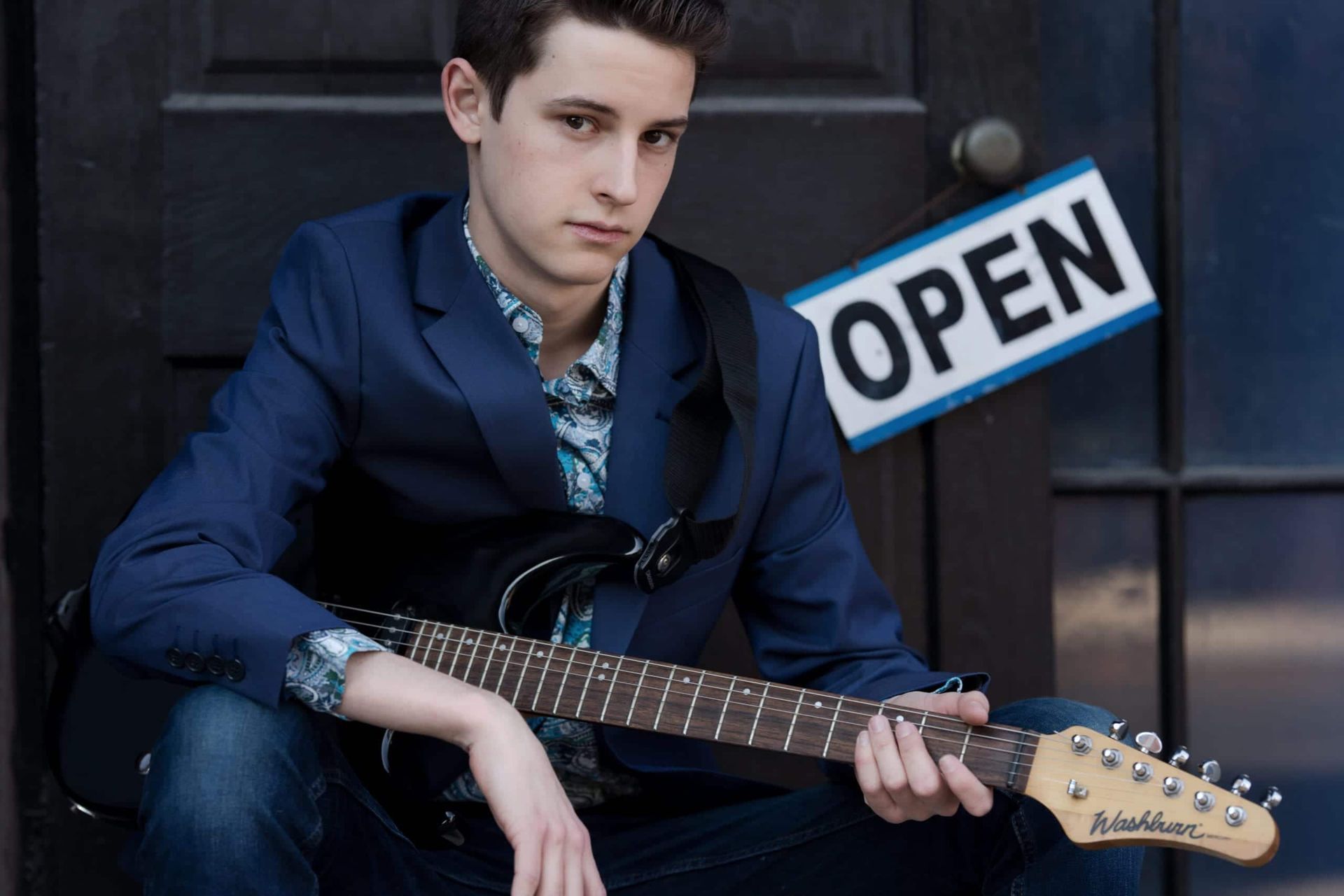 Young man seated, holding a guitar, wearing blue suit and patterned shirt, next to an 