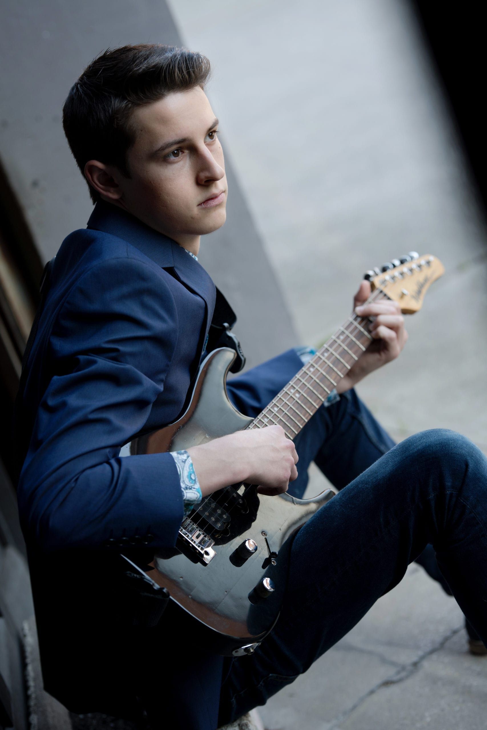 Young man in blue blazer playing electric guitar, seated outdoors, looking pensive.