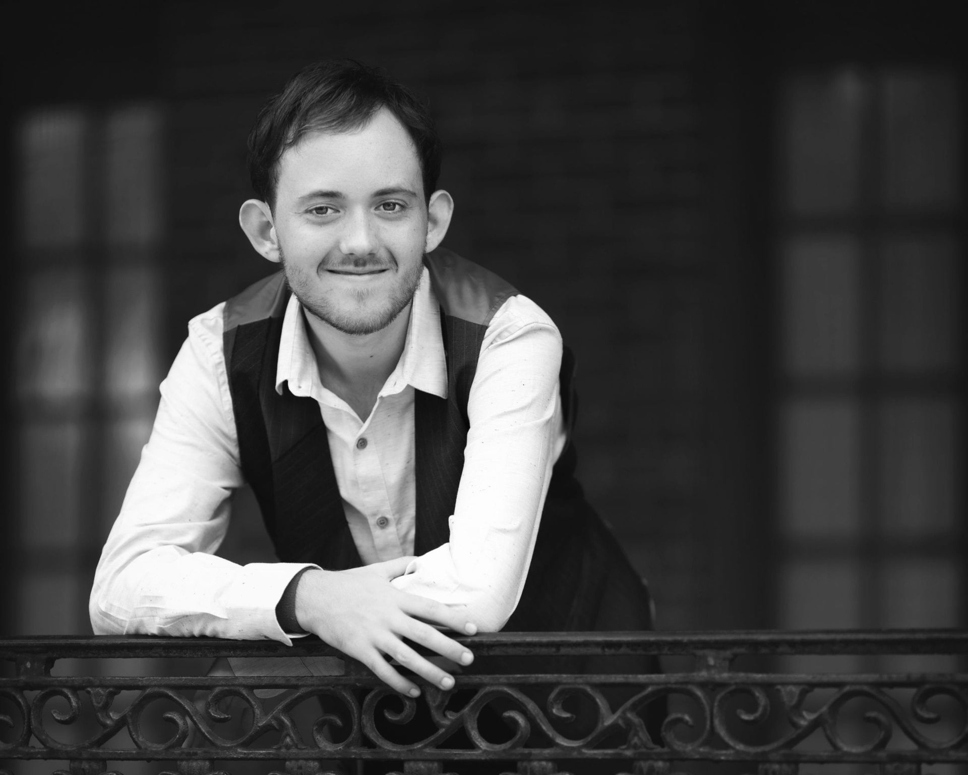 Man leaning on a wrought iron railing, smiling. Wearing a vest and button-down shirt. Balcony with a brick building in the background.