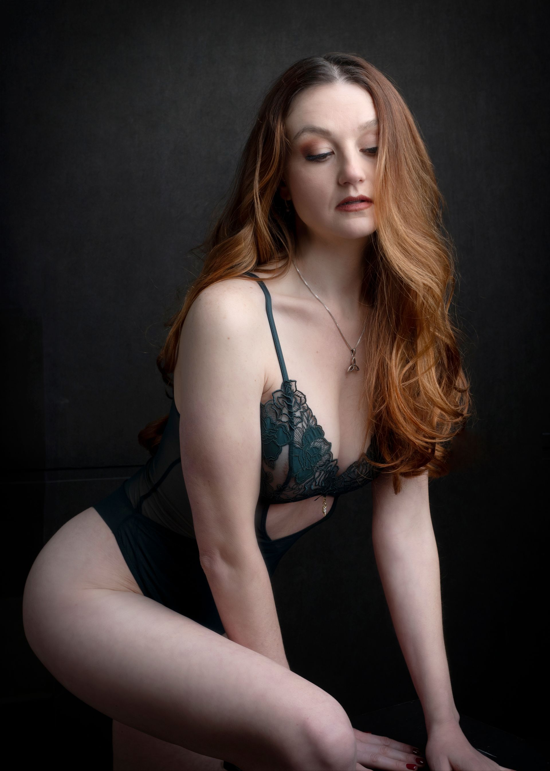 Woman with long red hair in a dark green lingerie set, leaning with a thoughtful expression. Black background.
