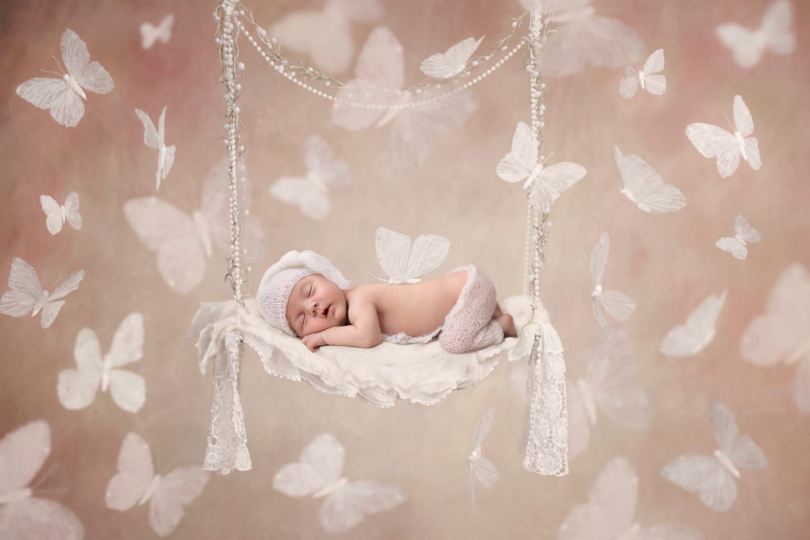Newborn baby asleep on a swing, surrounded by white butterflies on a pink background.