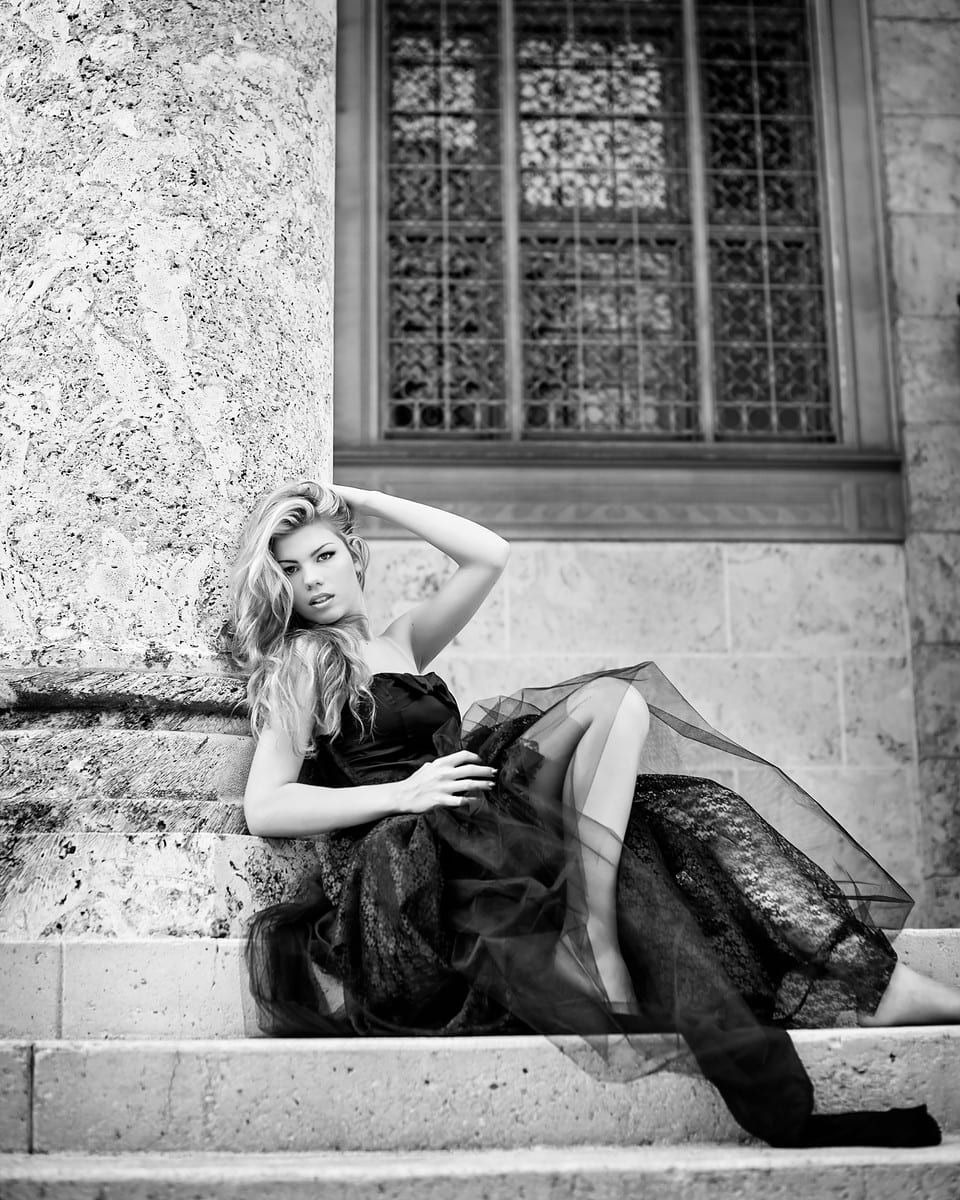 Woman in black dress reclines on stone steps, hand in hair, near window.