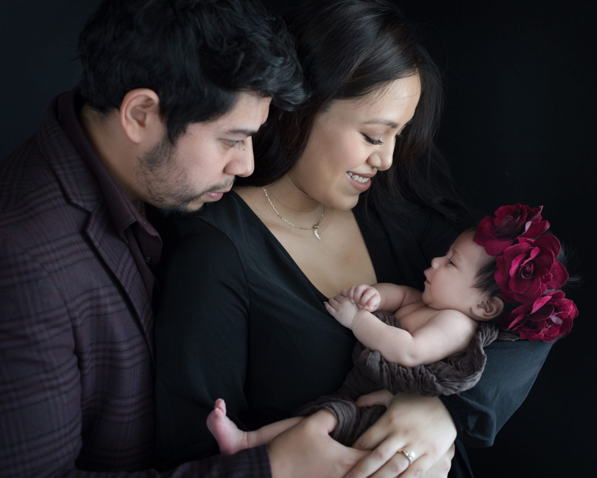 Parents gaze at newborn baby held in mother's arms; baby wears flower headpiece.