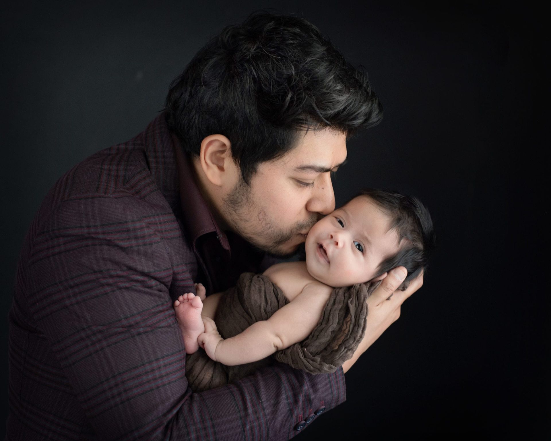 Father kisses newborn baby wrapped in a brown blanket; black background.