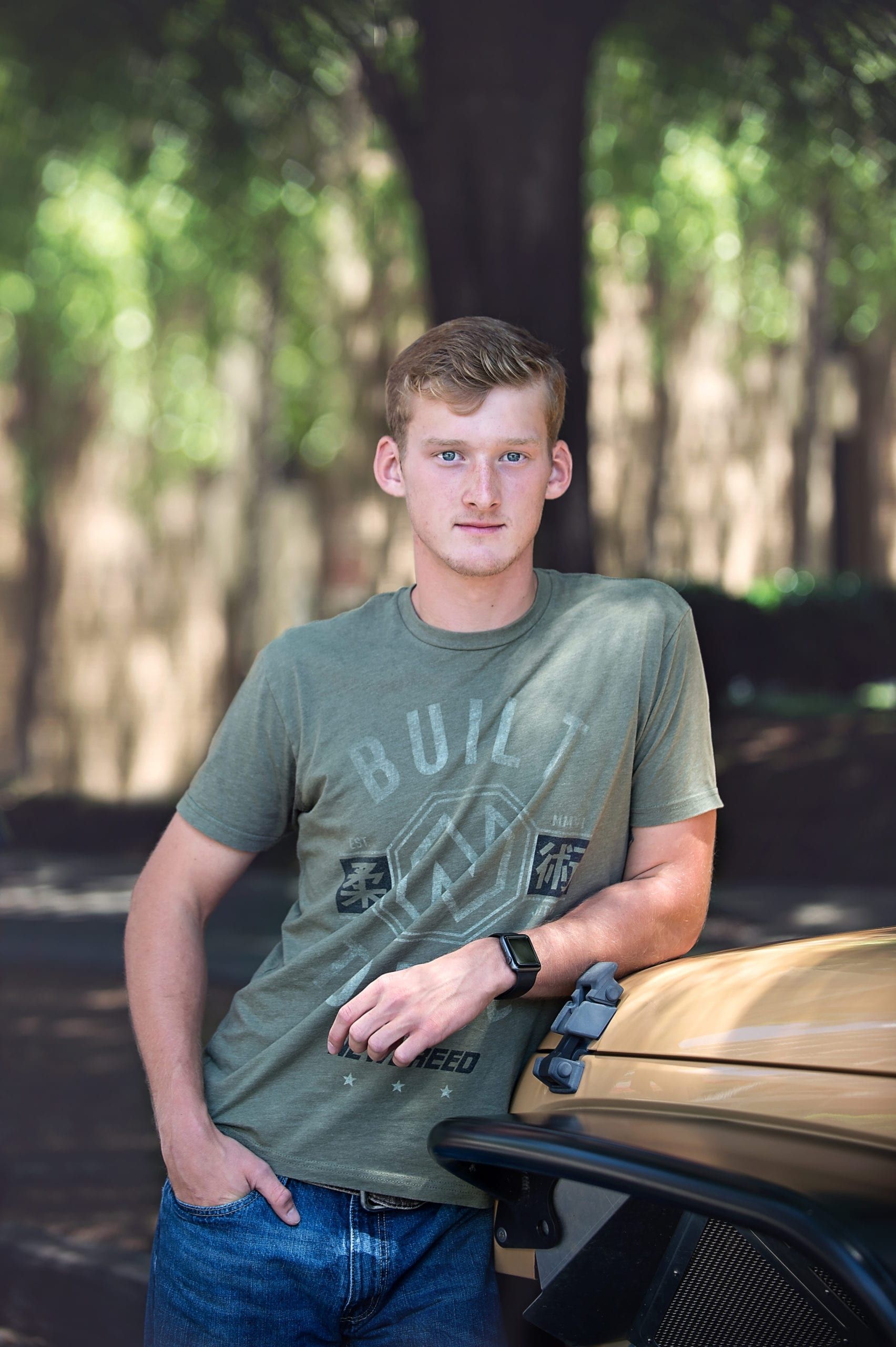 Young man in green shirt leaning against a vehicle, hand in pocket. Outdoors, trees in background.