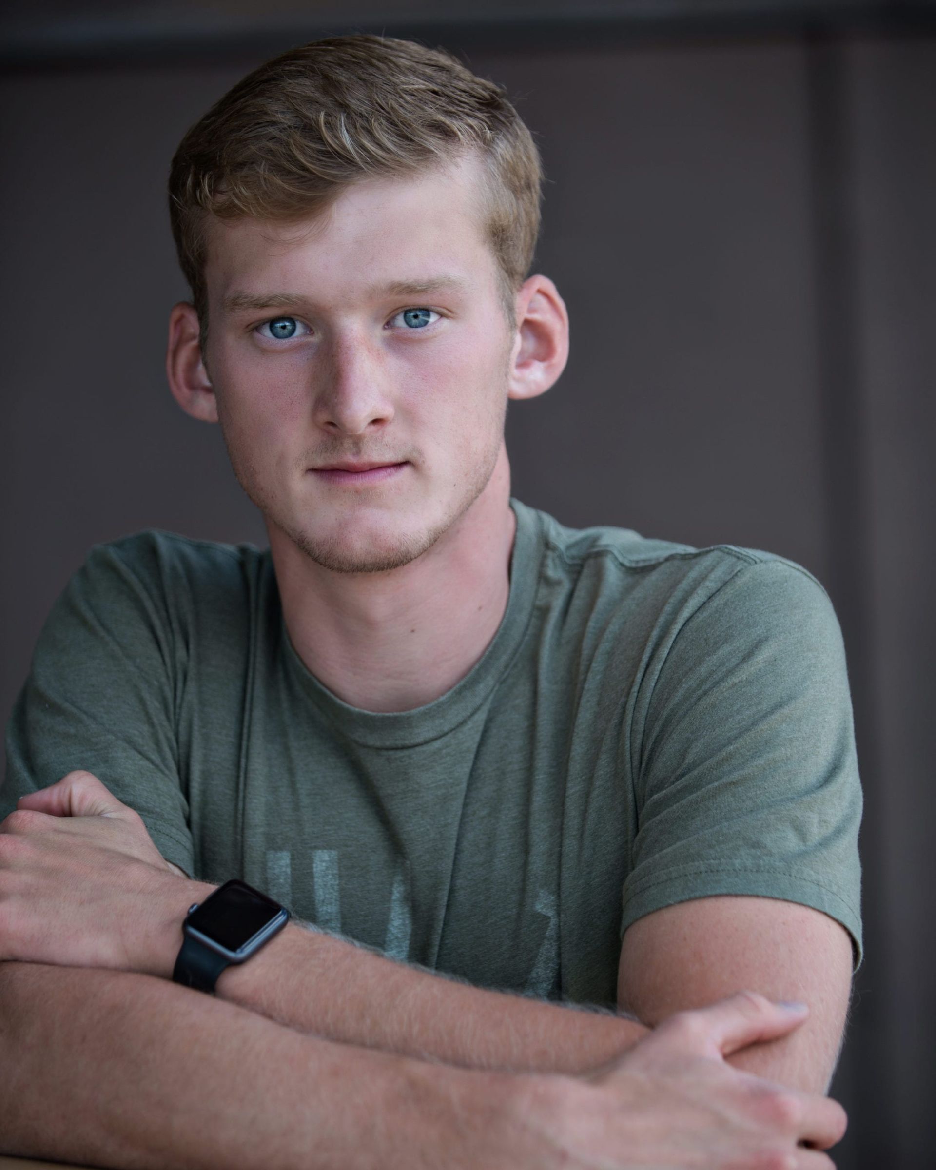 Young man with blonde hair and blue eyes, wearing a green shirt and smartwatch, arms crossed, neutral expression.