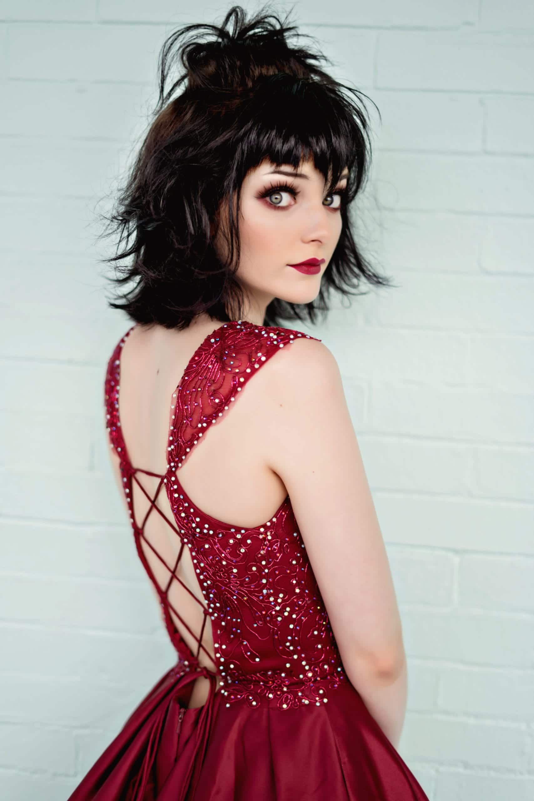 Woman with dark hair in a red formal dress, looking over her shoulder, standing in front of a white brick wall.