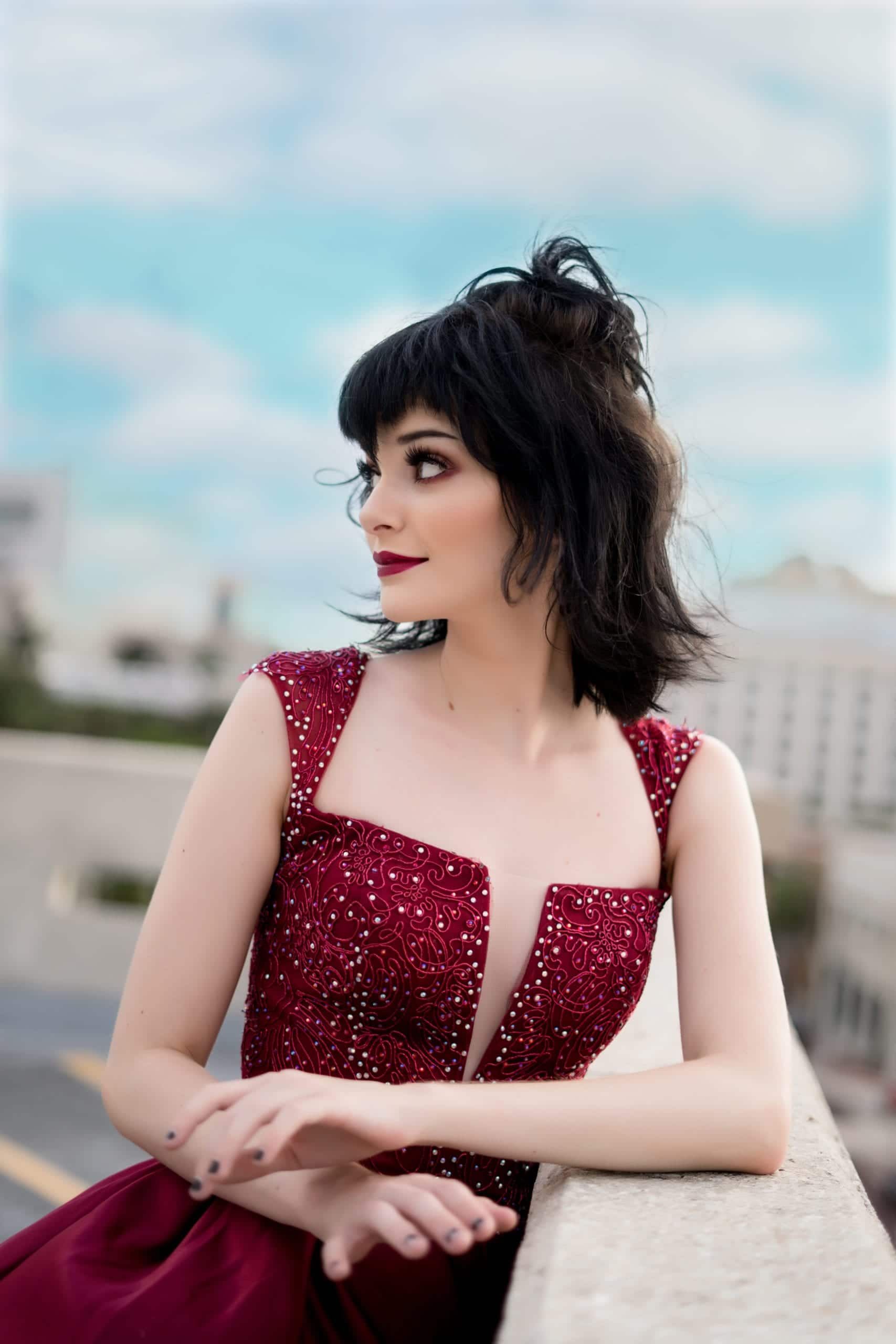 Woman in a burgundy beaded gown looks over her shoulder, leaning on a ledge.