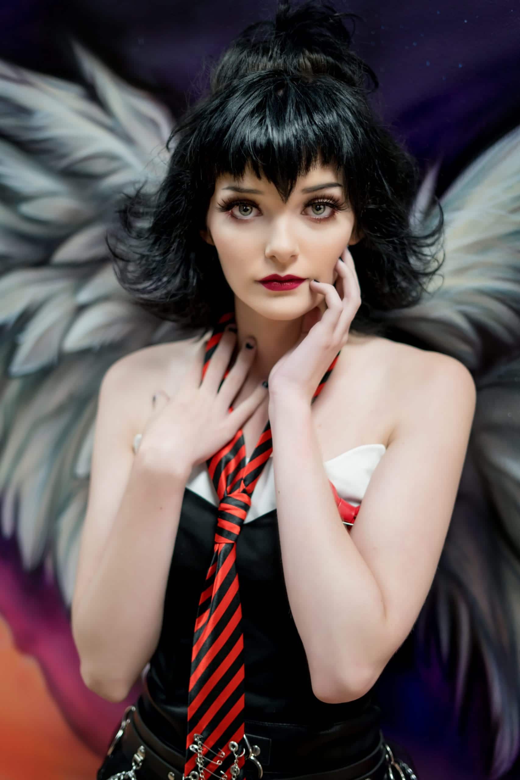 Woman with black hair and angel wings, red tie, hand near face, black and white outfit.