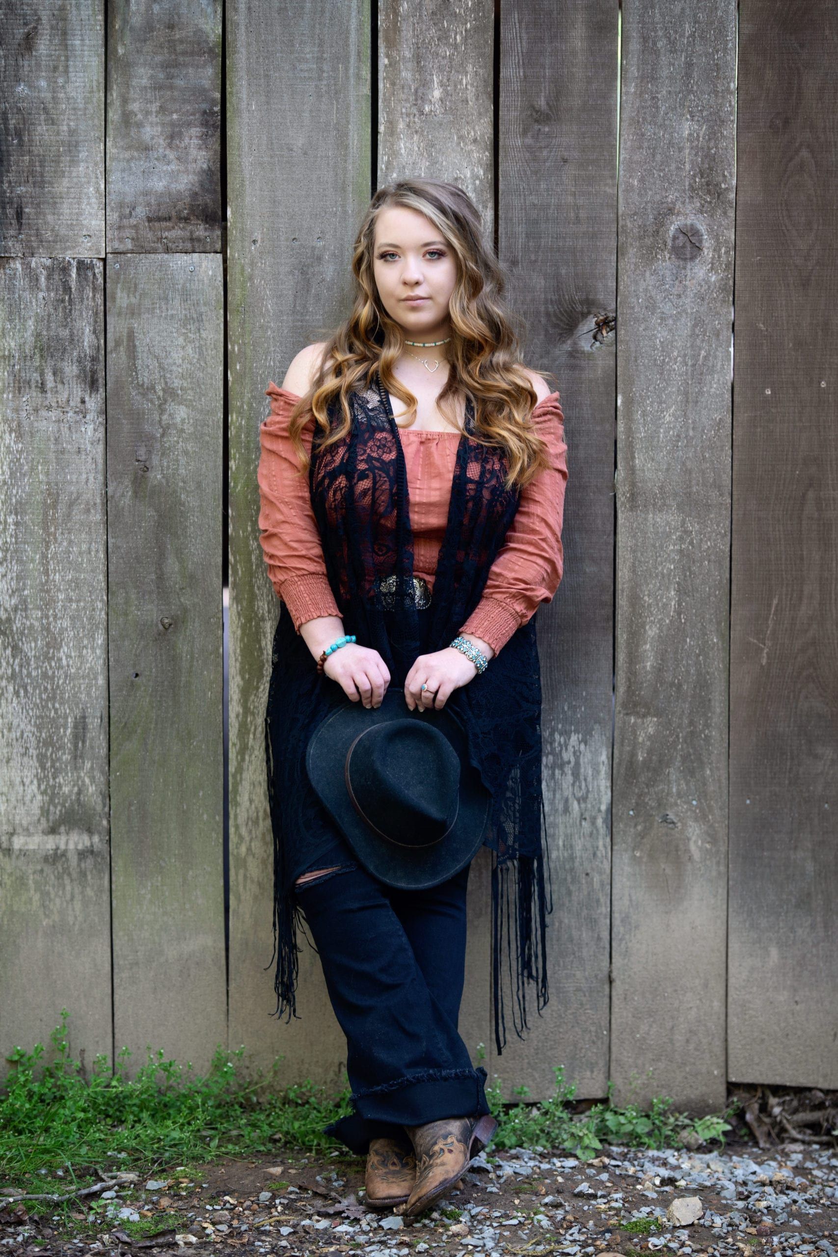 Woman in rust-colored top and fringed vest, holding a hat, stands against a weathered wooden wall.