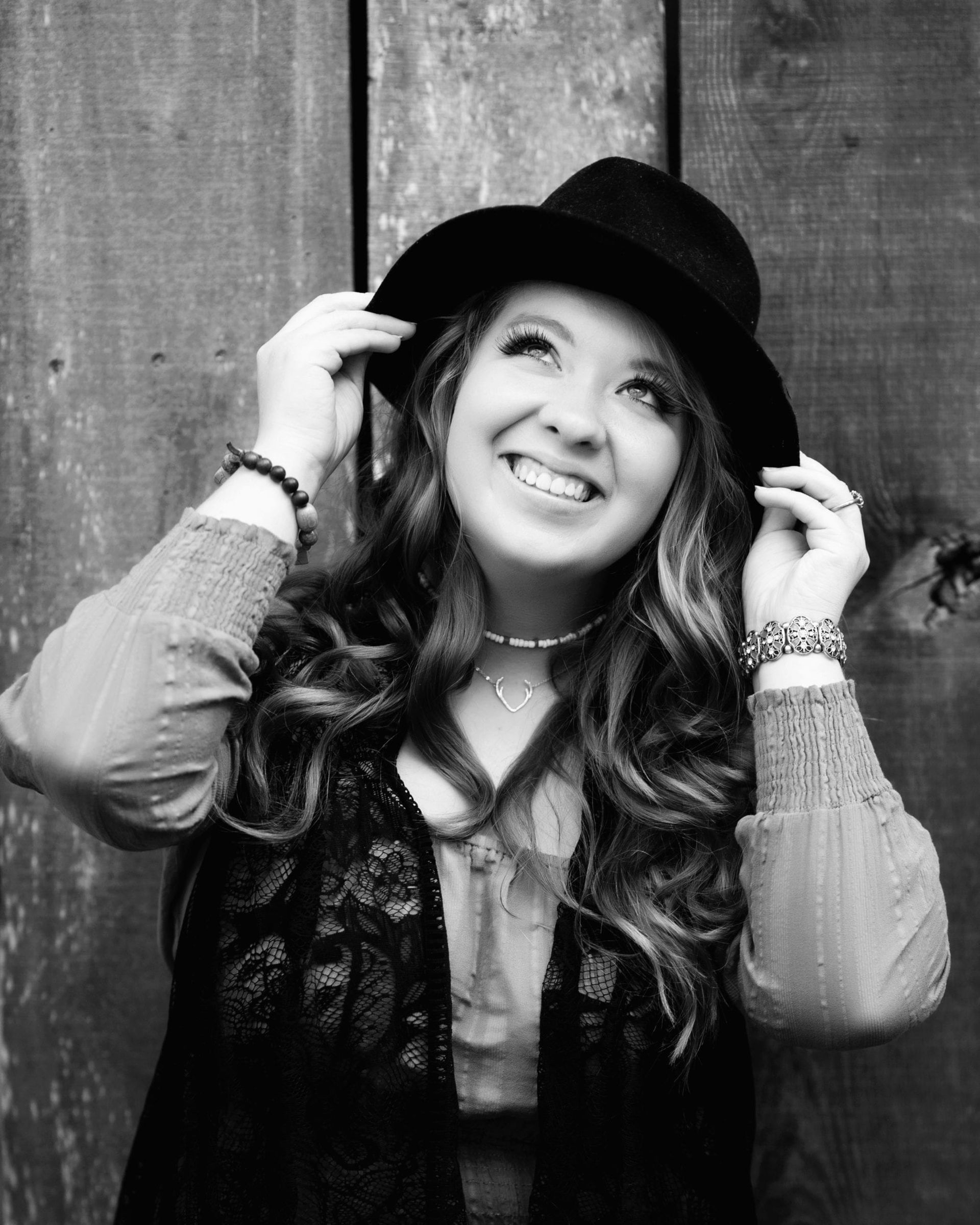 Woman in a hat smiles, leaning against a wooden wall.