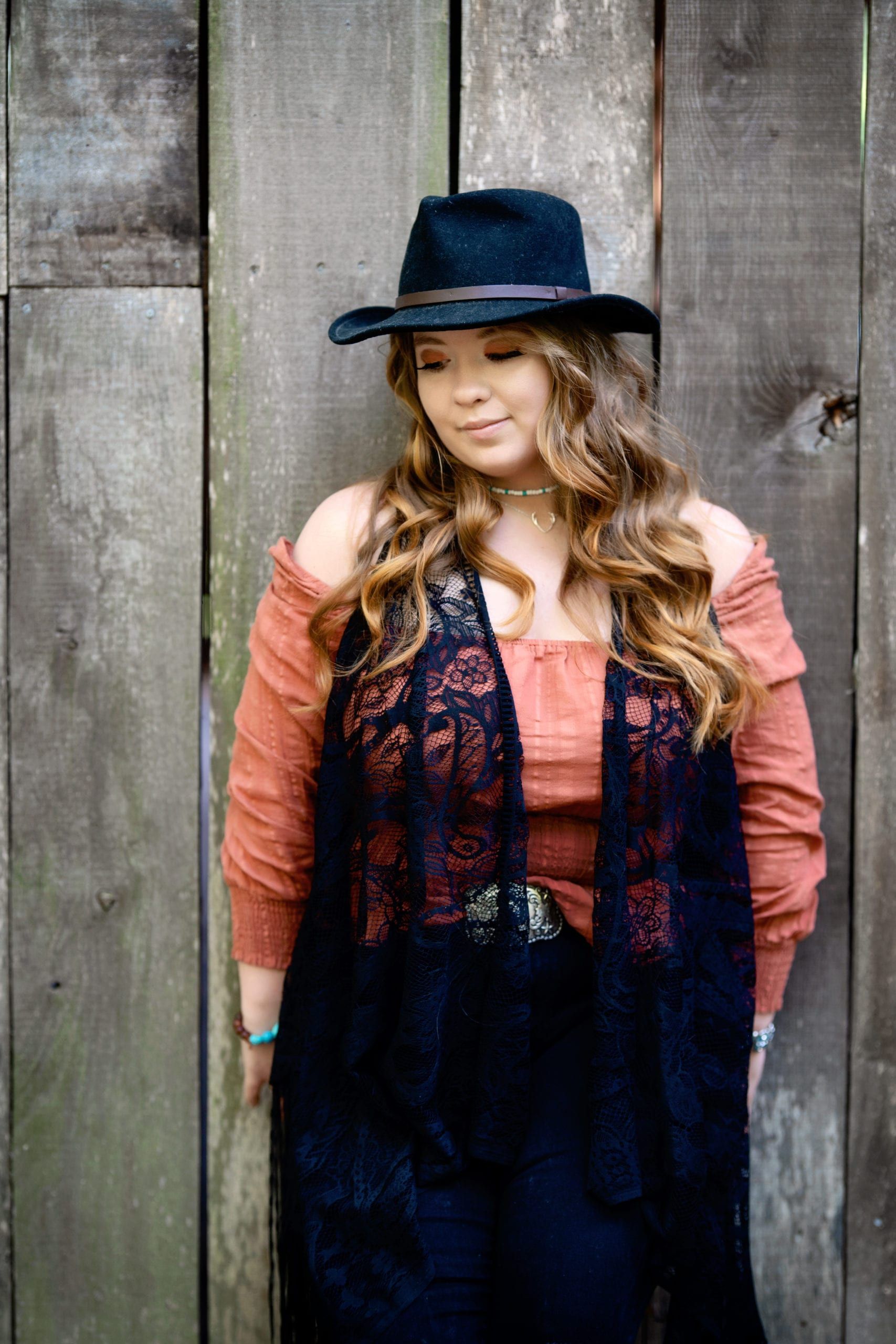 Woman in hat, leaning against a wooden wall. Wearing a long black vest over a rust-colored top.