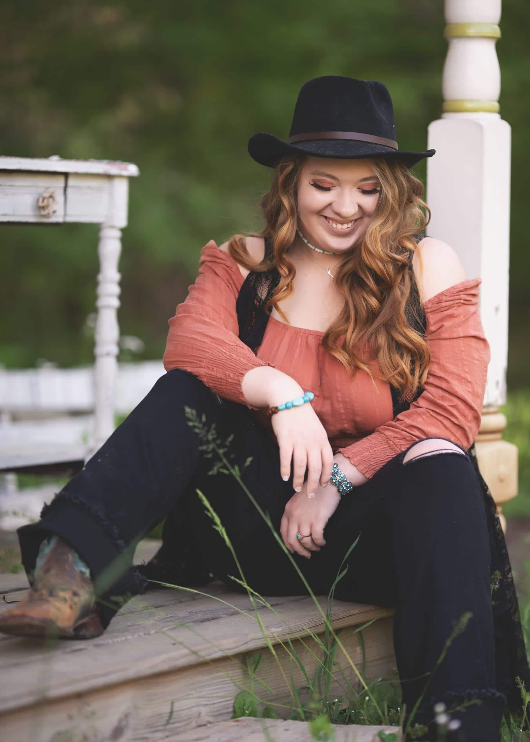 Woman in a cowboy hat and boots sits on steps, smiling. Wearing orange top and black pants.
