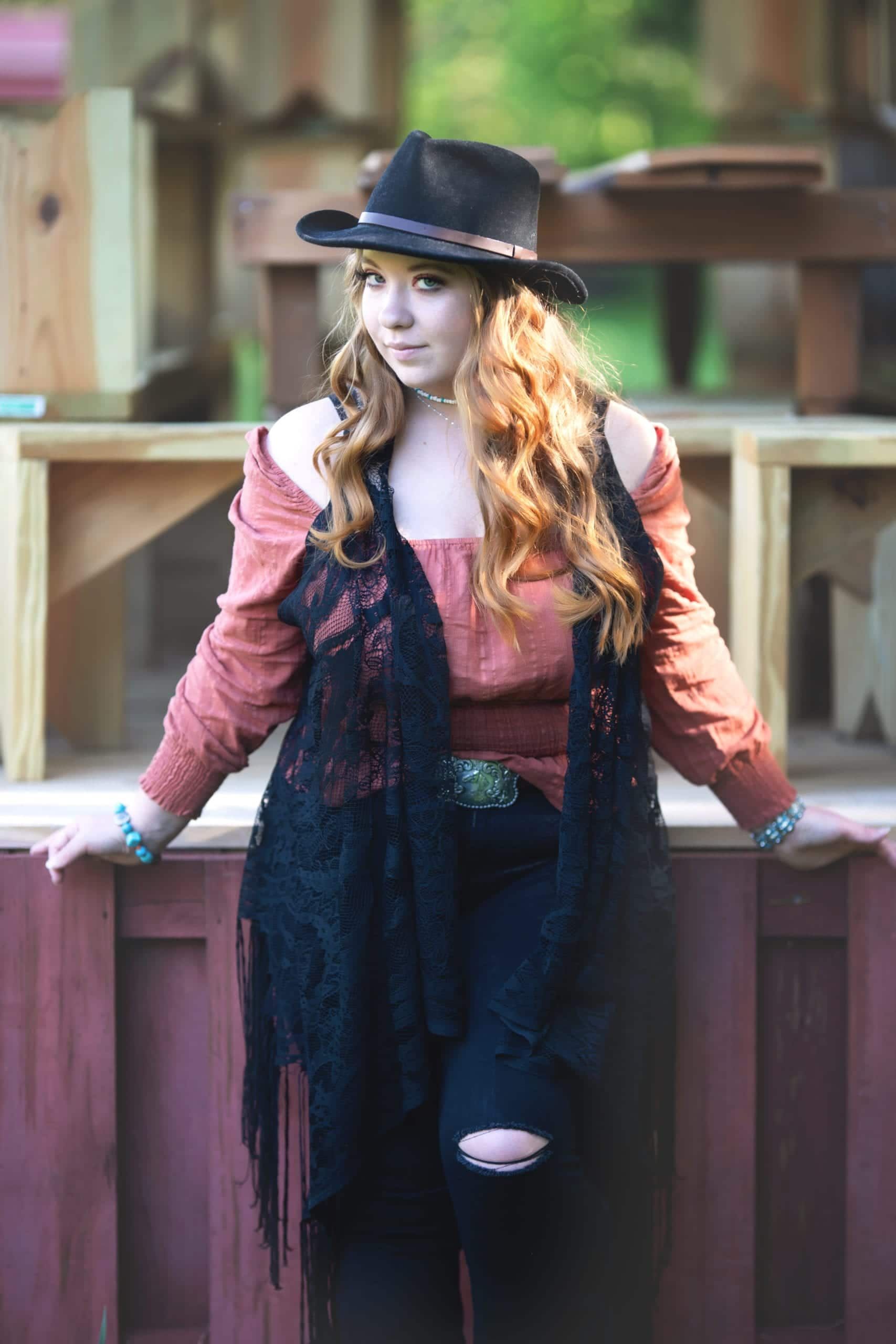Woman in cowboy hat, black vest, and ripped jeans leans on a wooden structure.