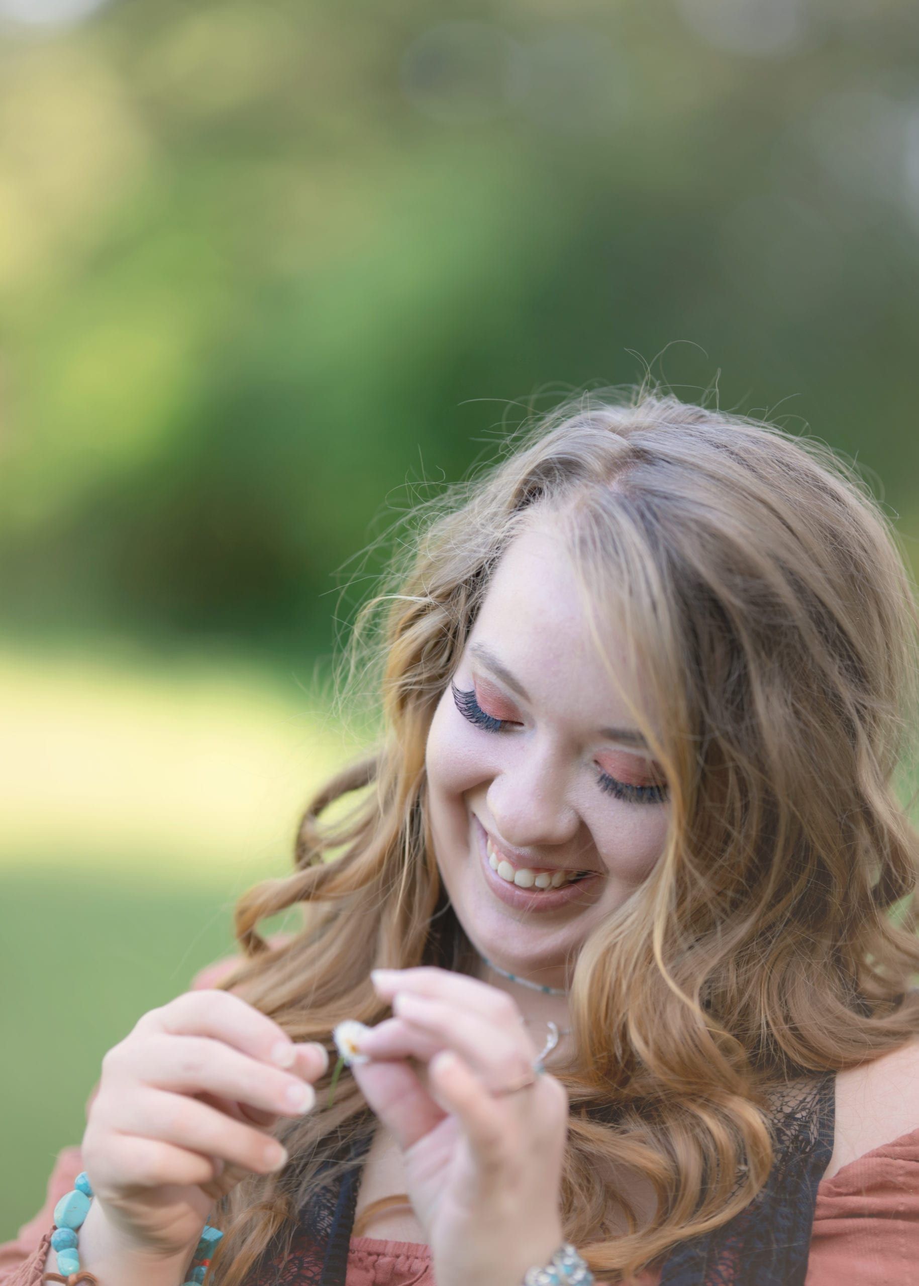 Woman with wavy blonde hair smiles, holding something small outdoors, green background.