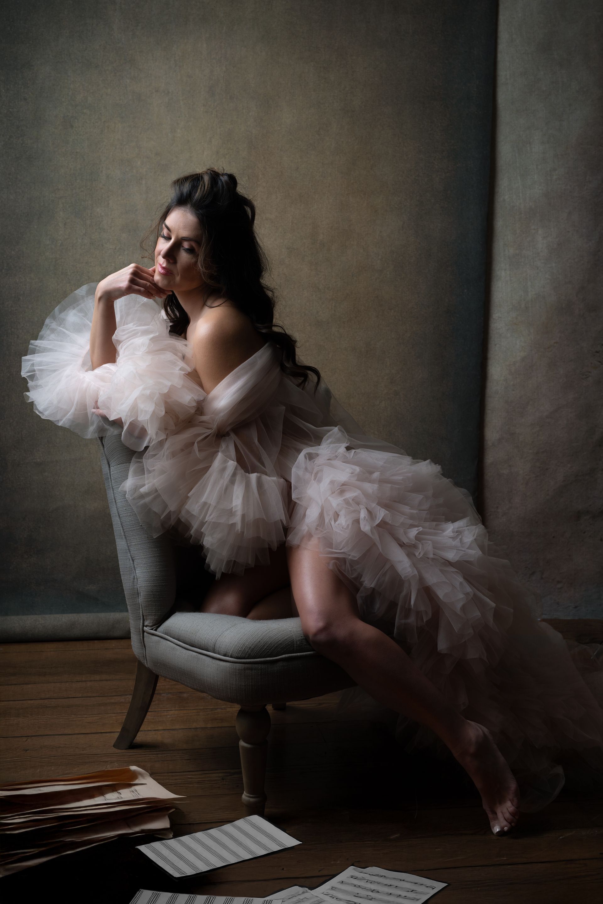 Woman in sheer, ruffled robe, sitting in an armchair, papers on the floor, against a mottled backdrop.