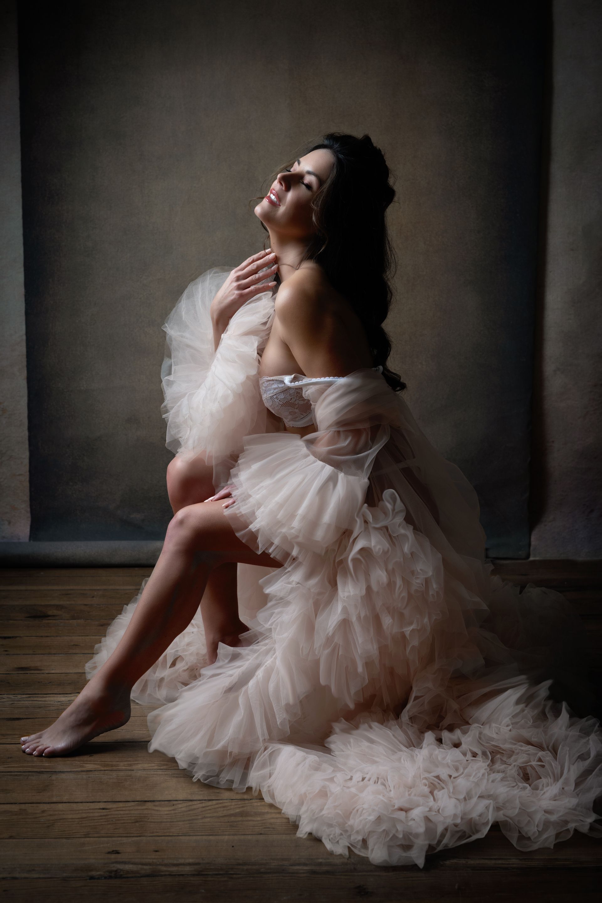 Woman in a fluffy, light-colored robe, seated on wooden floor, head tilted back, eyes closed, lit dramatically.
