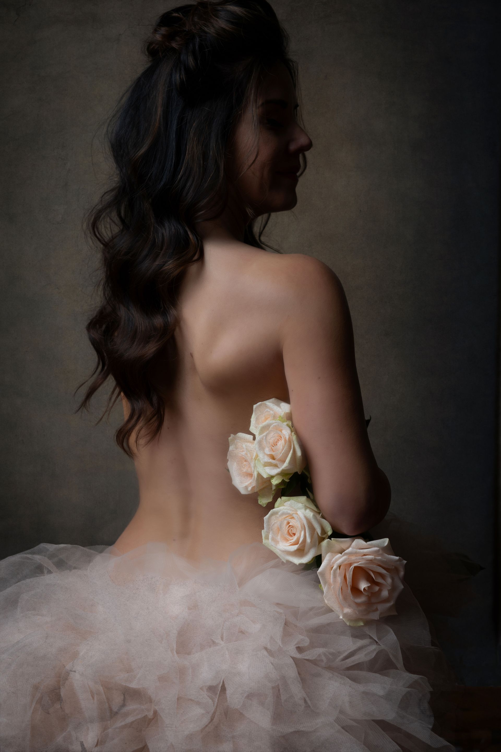 Woman, back to camera, bare shoulders, holding roses, tulle skirt. Dark background.